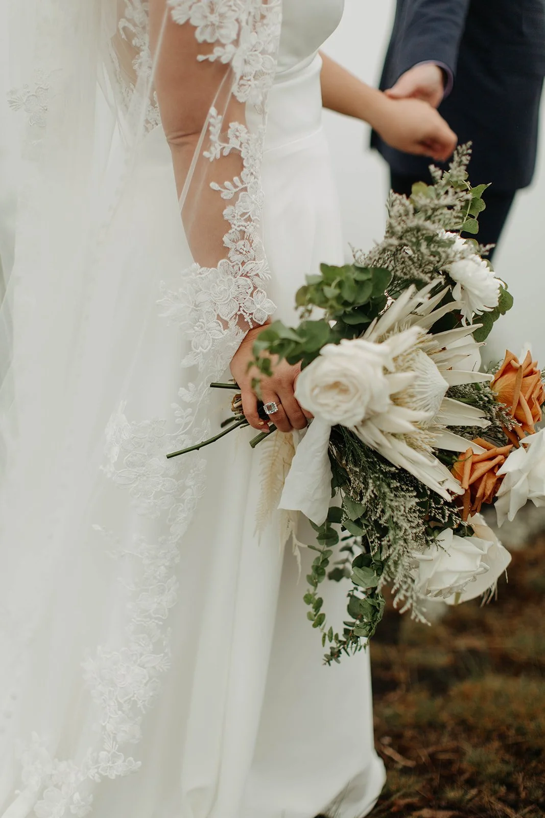 Close-up of a bride in a lace veil holding a white bouquet with greenery and dried floral accents.