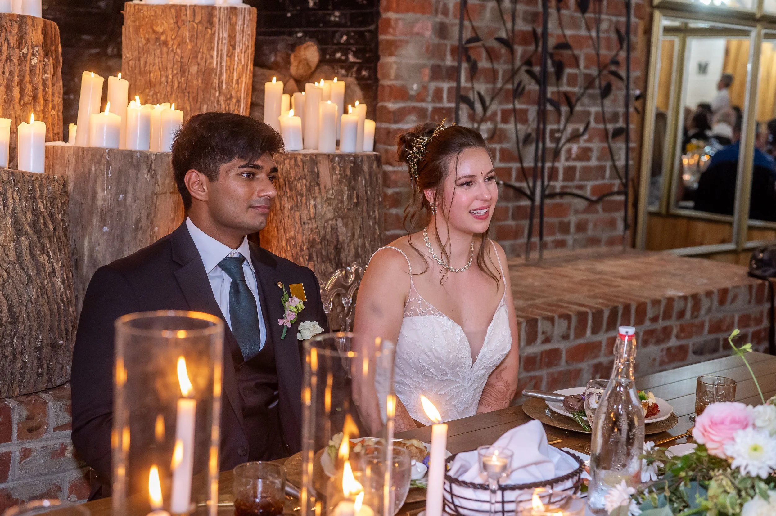 Bride and groom sit at a candlelit head table during dinner, listening to speeches in a rustic brick-and-wood venue.