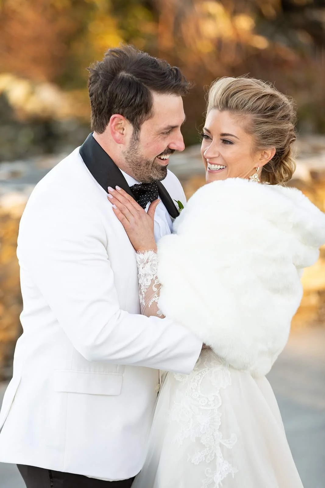 Winter wedding portrait of the couple smiling, groom in a white tuxedo and bride in a fur wrap.