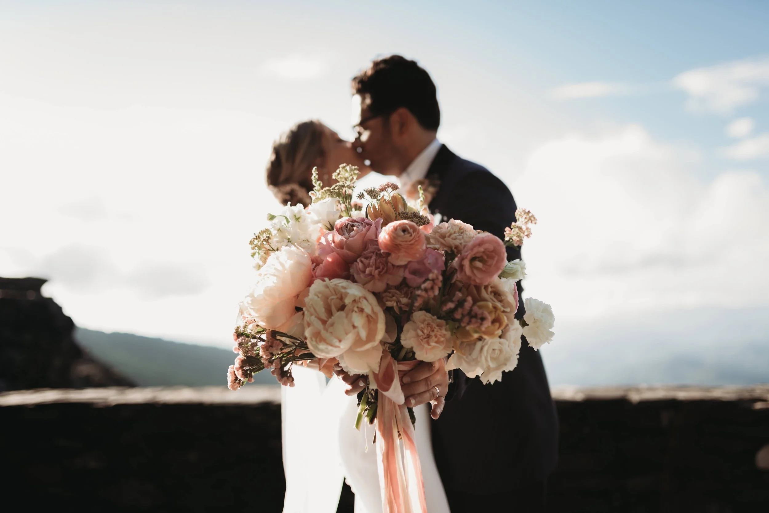 Blush and ivory bridal bouquet in focus while the bride and groom kiss in the background at a mountain overlook.