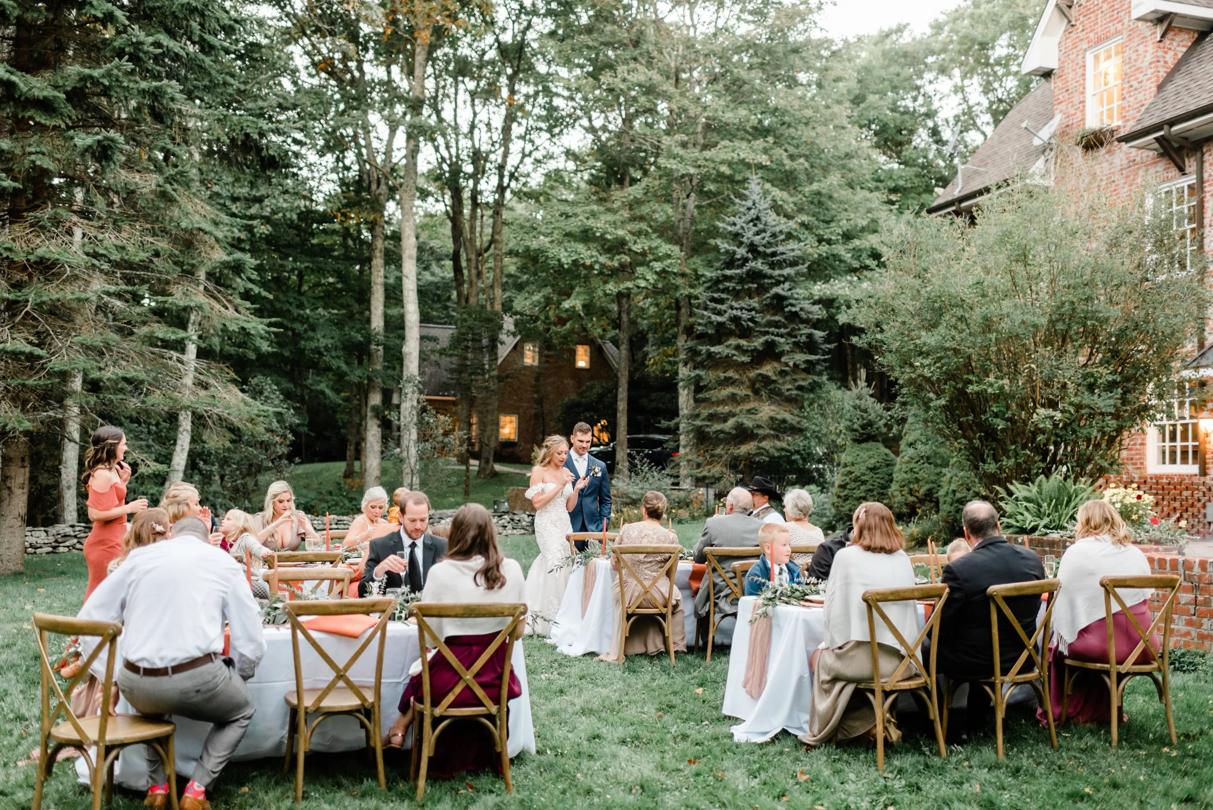 Intimate garden wedding reception with guests seated at round tables, wooden chairs, and greenery outside a brick estate.