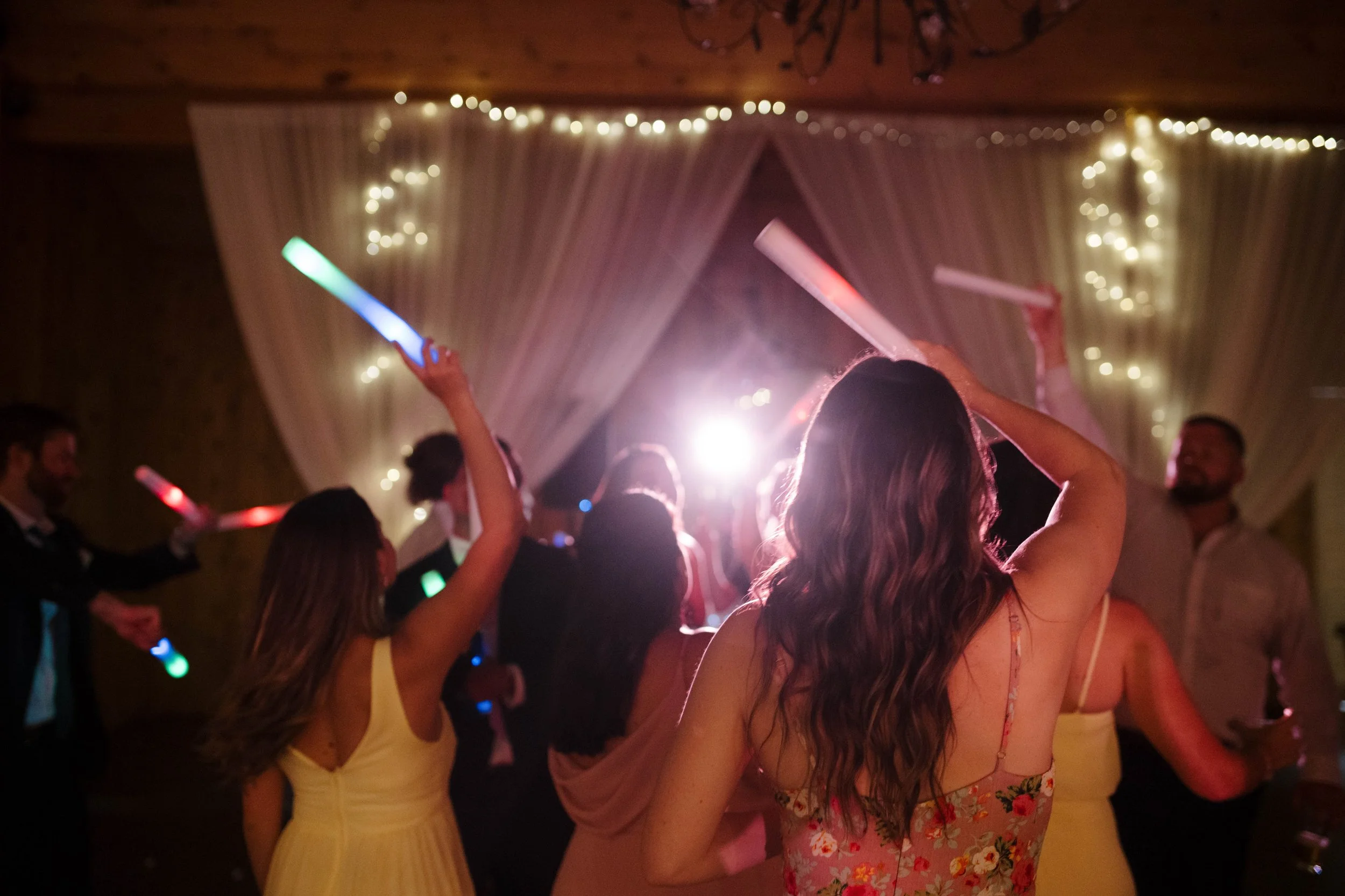 Wedding guests dancing with colorful glow sticks during a lively evening reception with string lights and white draping.
