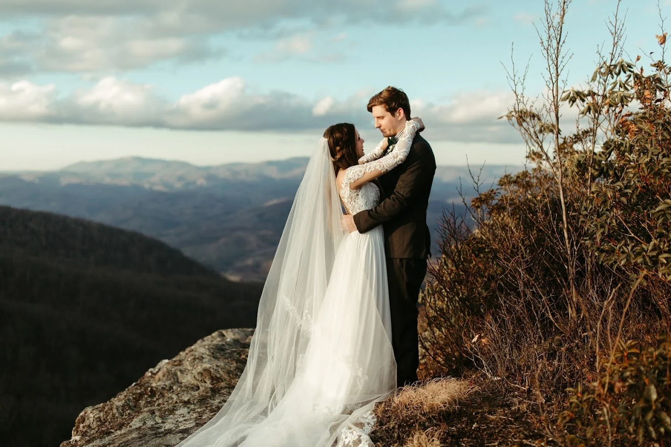 Newlyweds embracing on a mountain cliff overlook with the bride’s long veil trailing over the rocks.