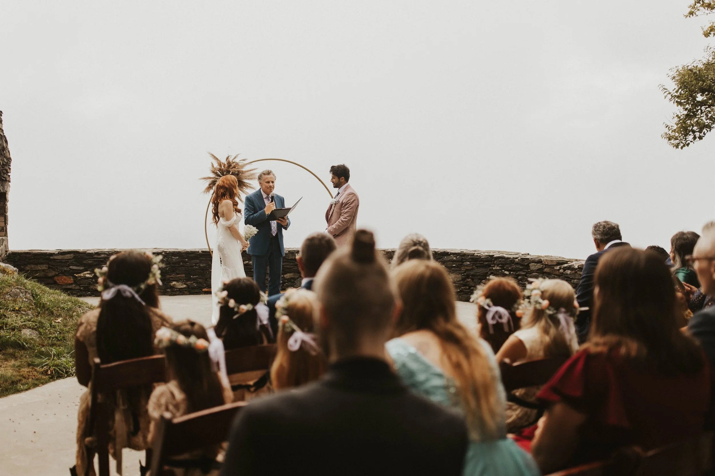 Intimate mountain wedding ceremony with bride, groom, officiant, seated guests, and a circular floral arch overlooking a stone wall.