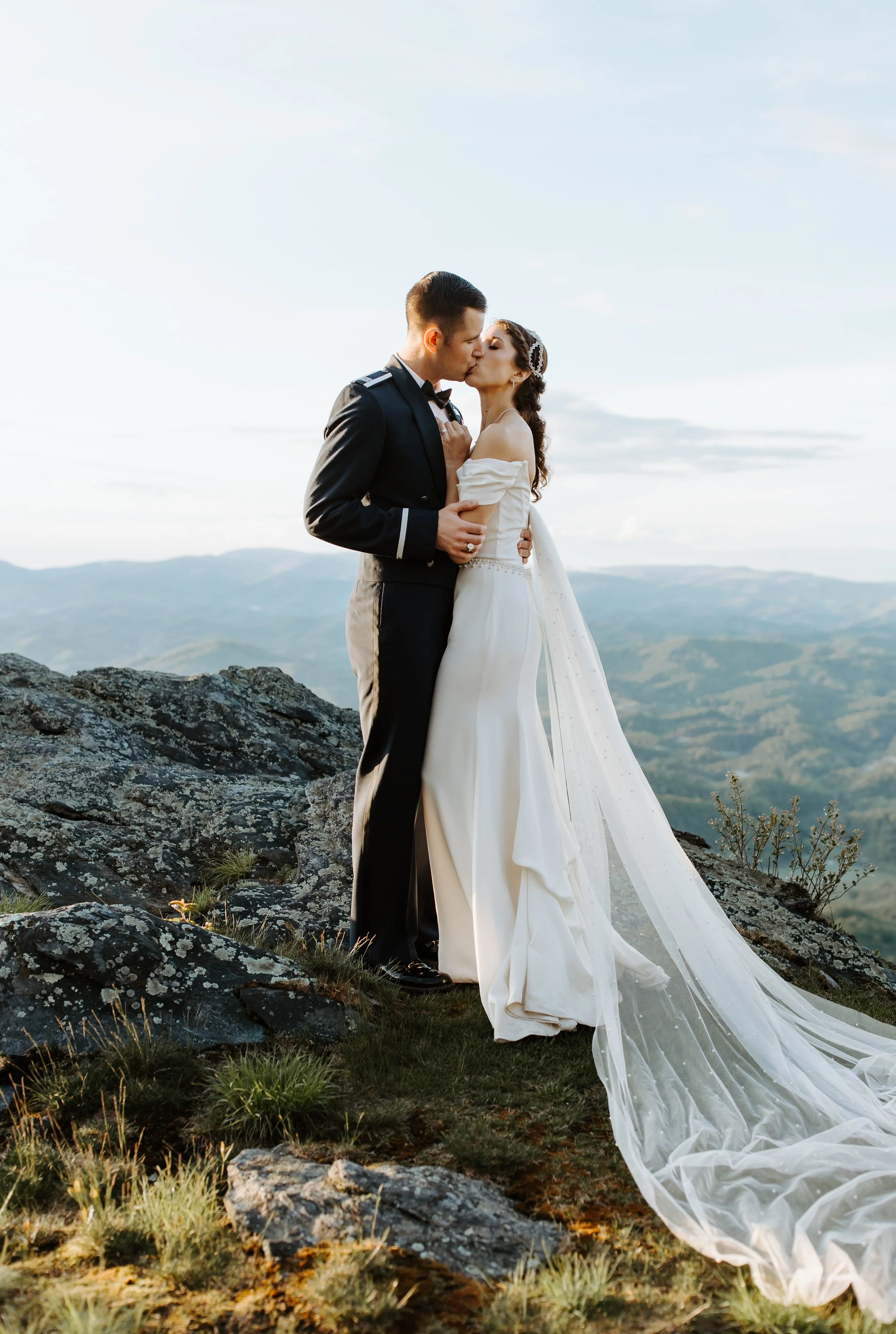 Bride and groom kissing on a mountaintop with sweeping views and a long veil flowing behind the bride.