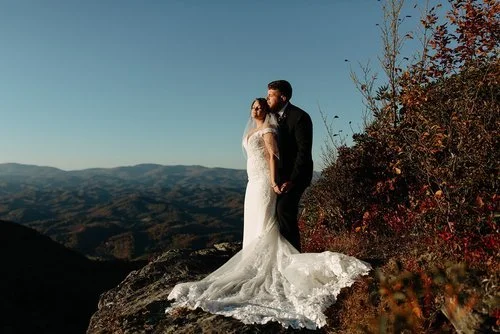 A bride and groom embrace on a rocky mountain, the bride's lace dress and veil flowing, with a scenic mountain view in the background.