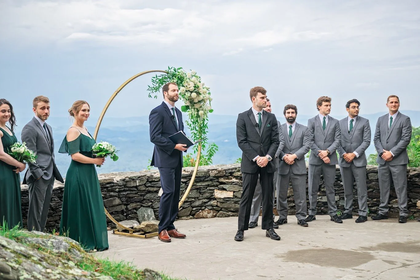 Wedding party standing on a mountain overlook beside a circular floral arch during an outdoor ceremony