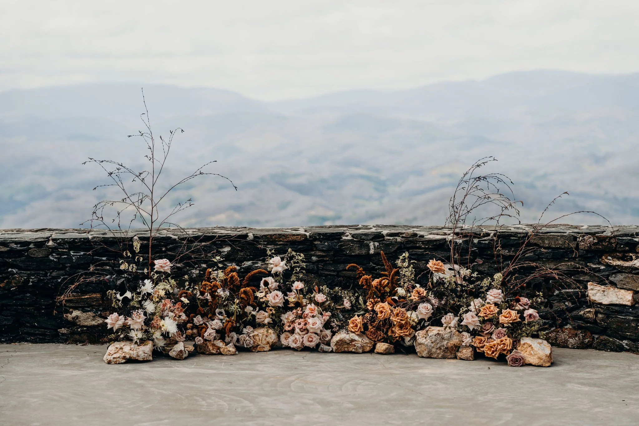 Low floral installation arranged along a stone wall at a mountain overlook ceremony site.