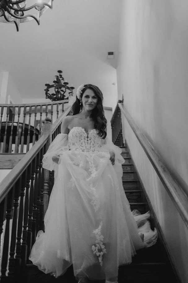Black-and-white portrait of a bride walking down a staircase in an off-the-shoulder wedding gown