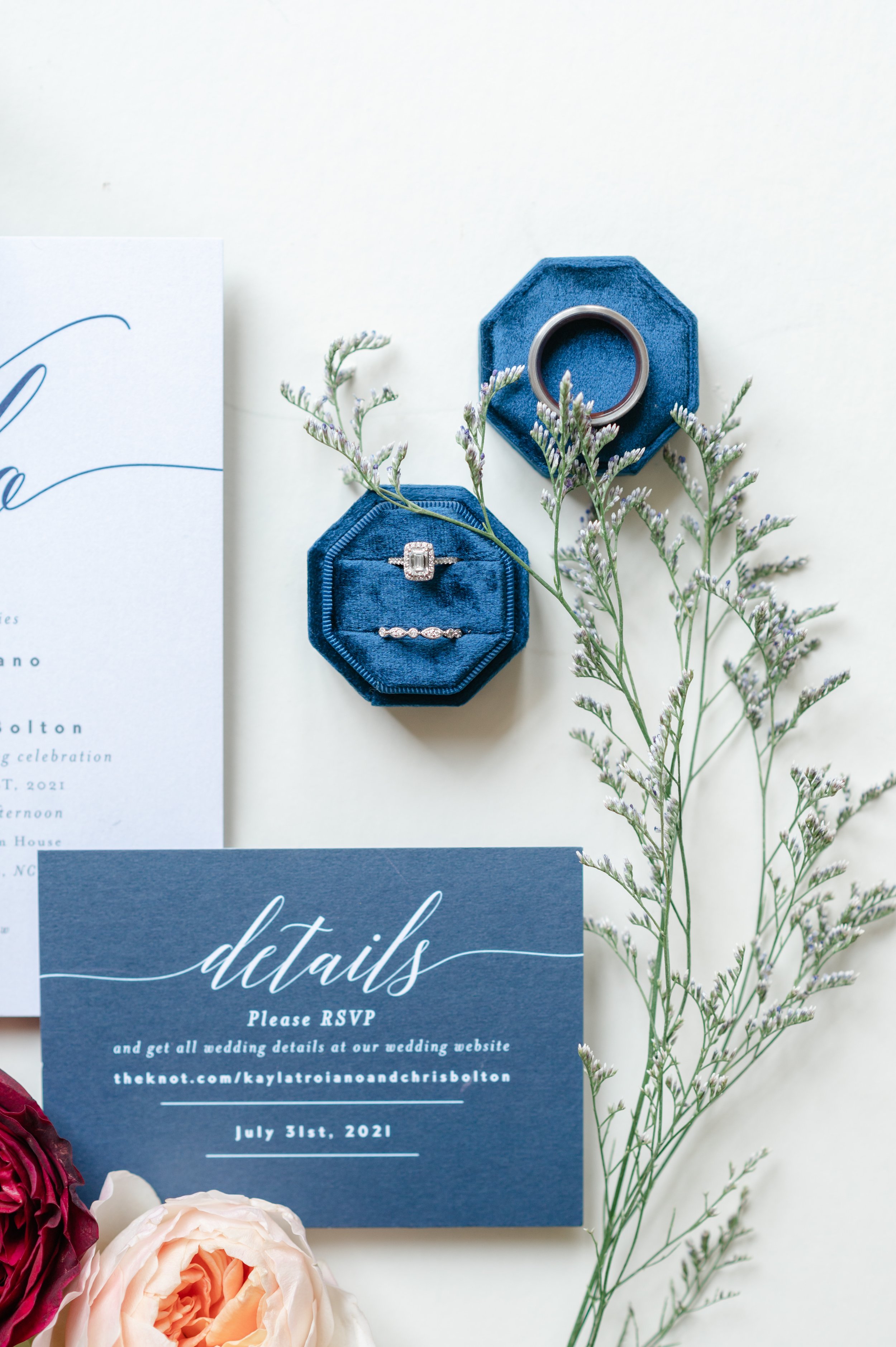 Wedding rings in a blue velvet ring box with greenery and a blue details card in a flatlay.