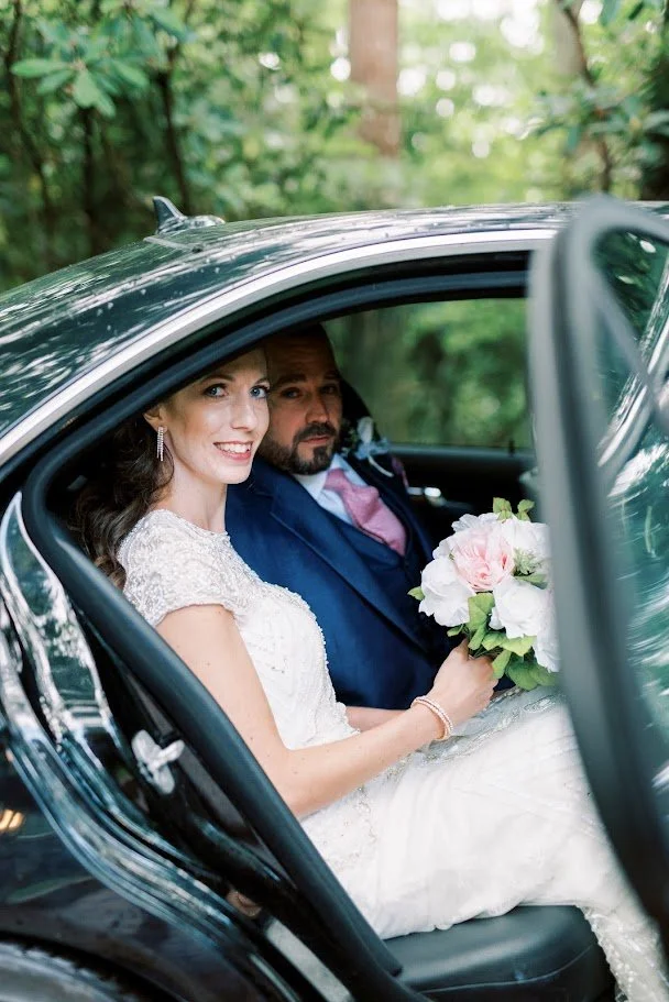 Bride and groom sitting in the back seat of a car on their wedding day, bride holding a pink-and-white bouquet
