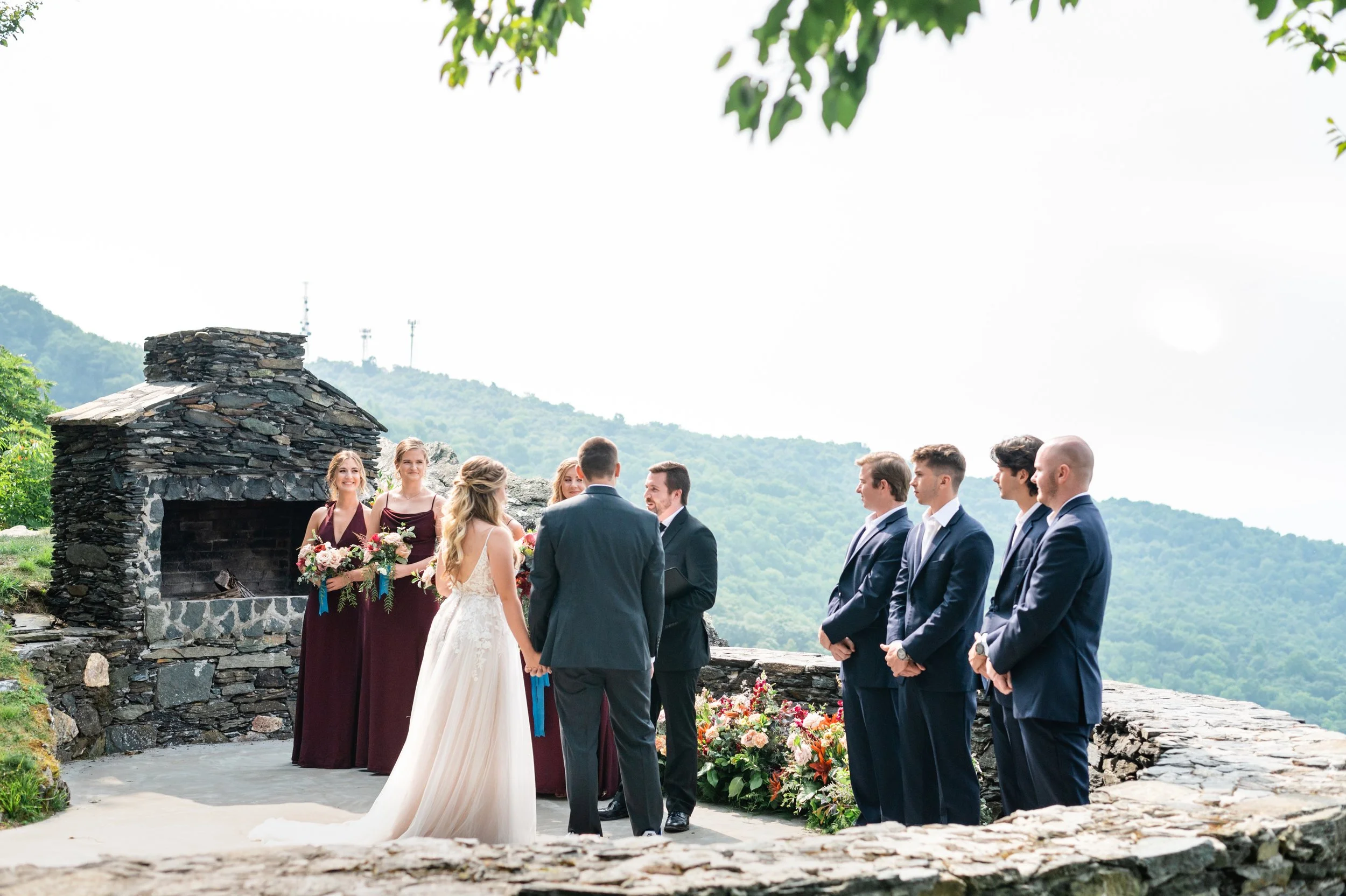 Couple standing with officiant at a mountain overlook ceremony beside a stone structure.