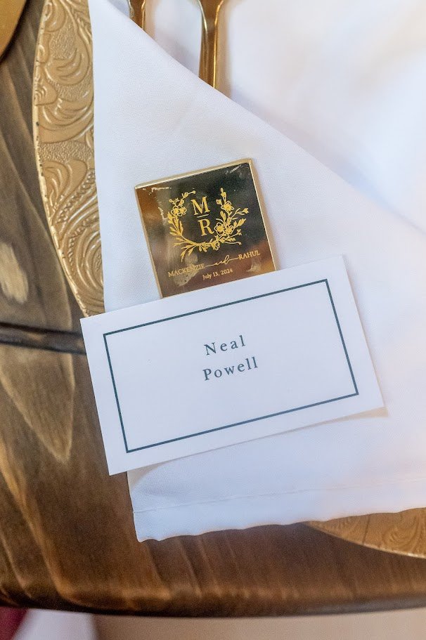 Close-up of a place card reading “Neal Powell” tucked into a folded white napkin on a gold charger.