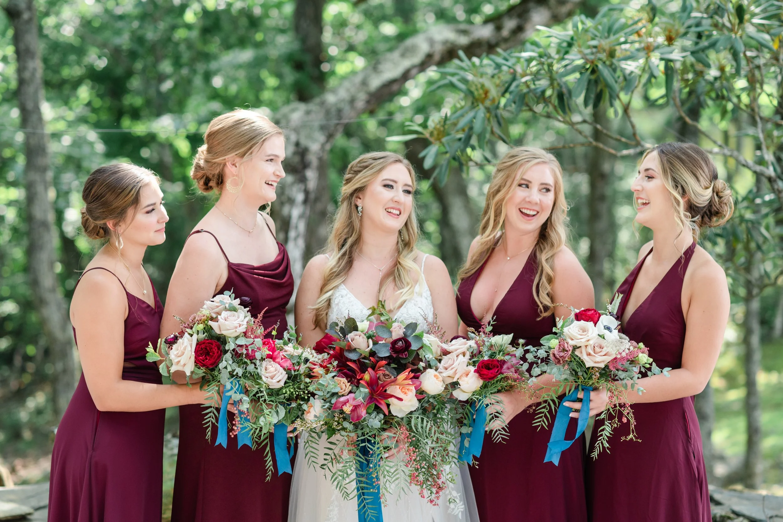 Bride with bridesmaids in burgundy dresses holding bouquets, smiling outdoors.