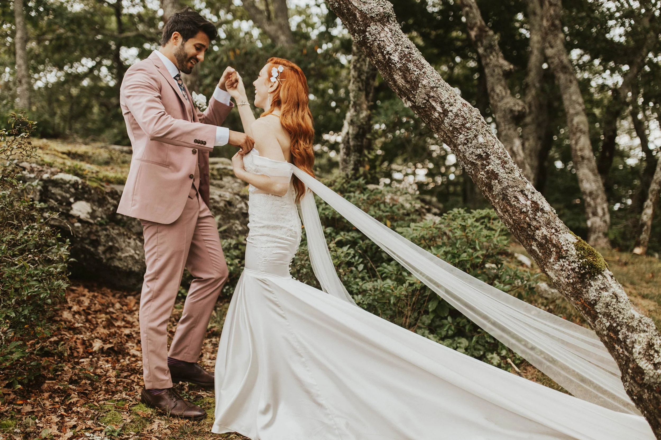 Bride and groom smiling during a romantic veil portrait in a wooded mountain setting with rocks, trees, and flowing white fabric.