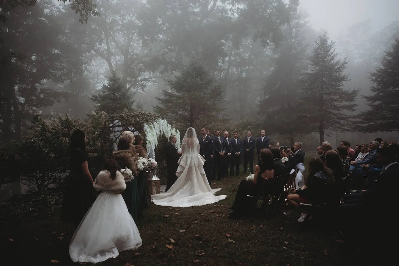 Wide view of a wedding ceremony with guests seated in a foggy forest and the couple at the altar.