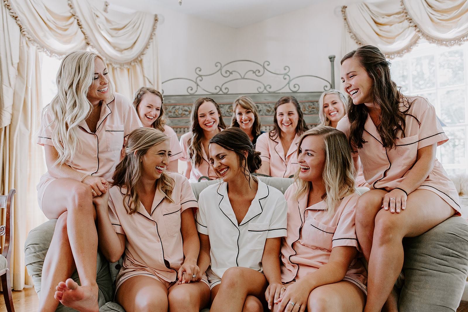 Bride and bridal party laughing together on a bed in matching pink getting-ready pajamas.