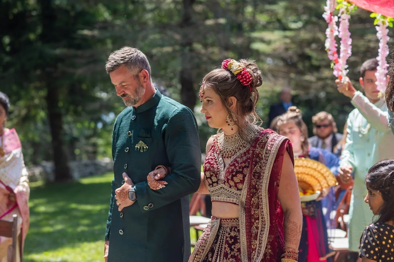 : Bride in embellished attire walks arm-in-arm with an older man outdoors as guests watch, sunlight filtering through trees.