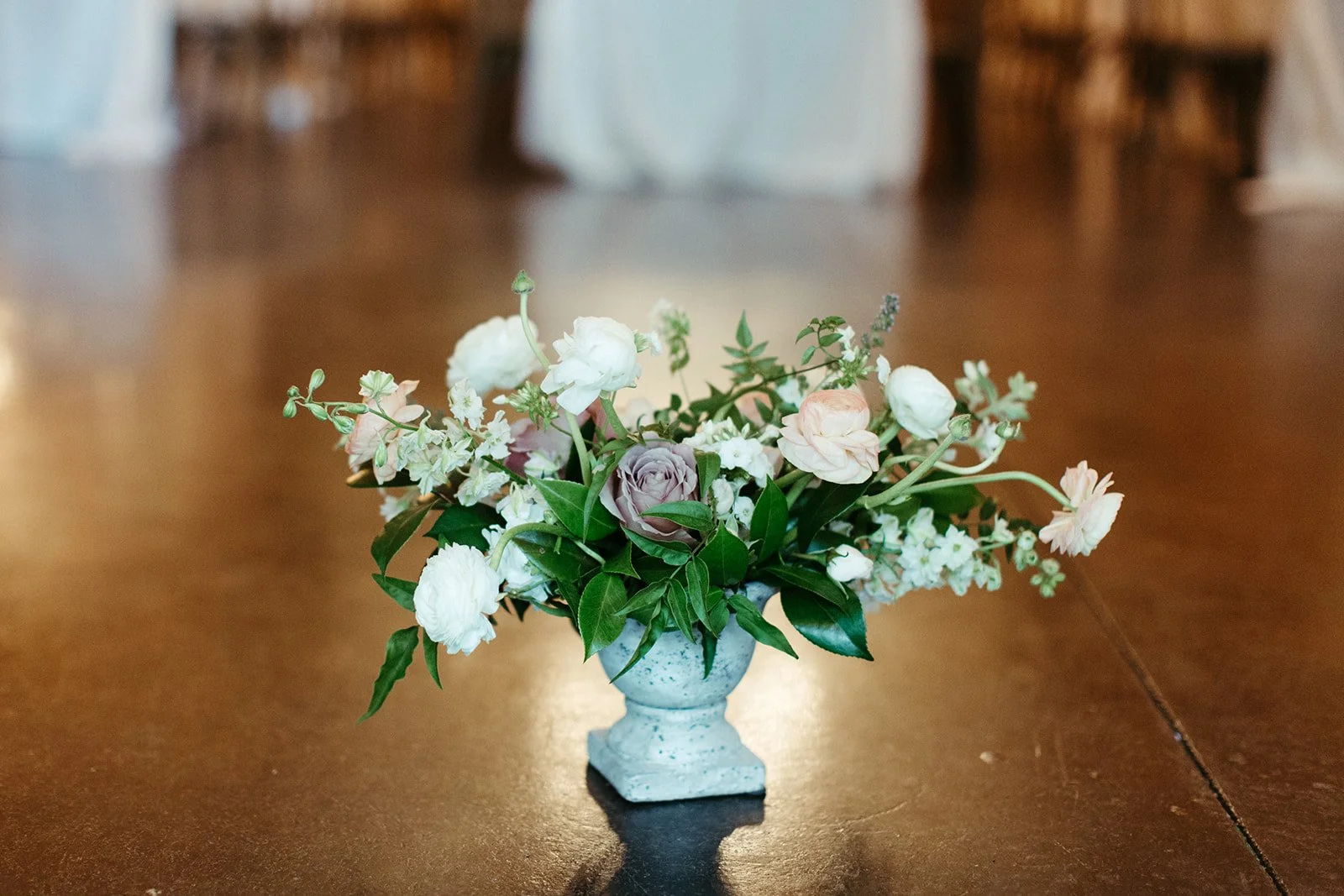 Low floral centerpiece with roses and ranunculus in a white compote vase on a wooden table.