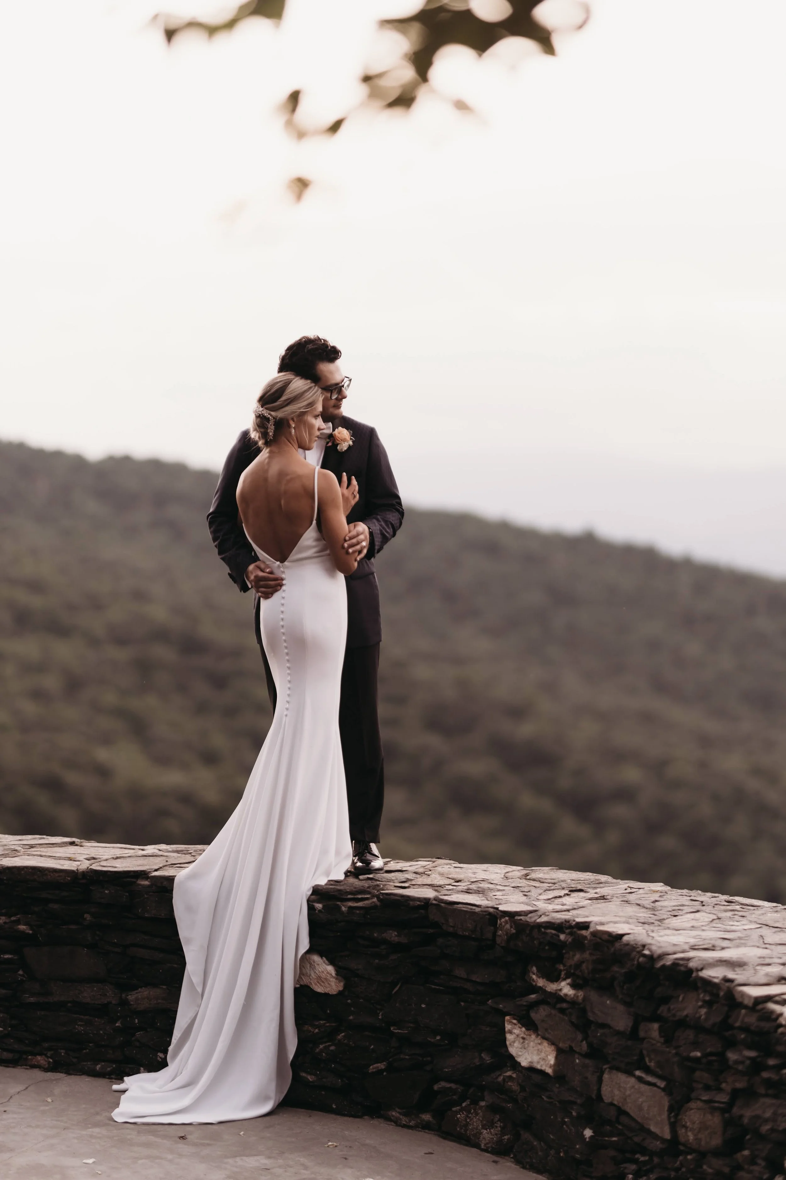 Bride and groom embracing on a stone wall at a scenic mountain overlook with the bride’s long train flowing behind her.