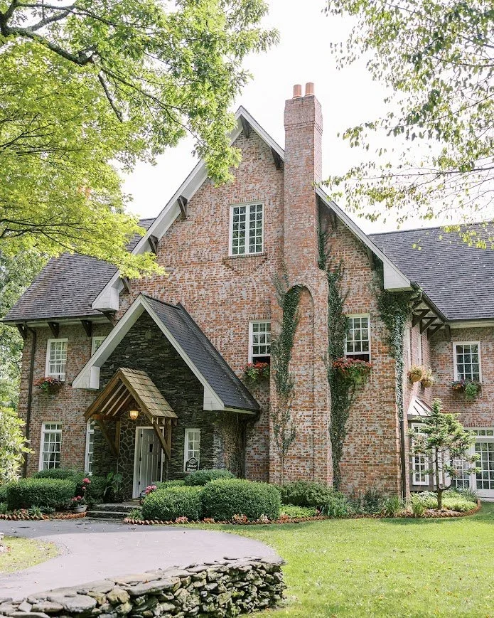 Front view of an ivy-trimmed brick manor with steep gables, a tall chimney, and lush landscaping along a curved drive.