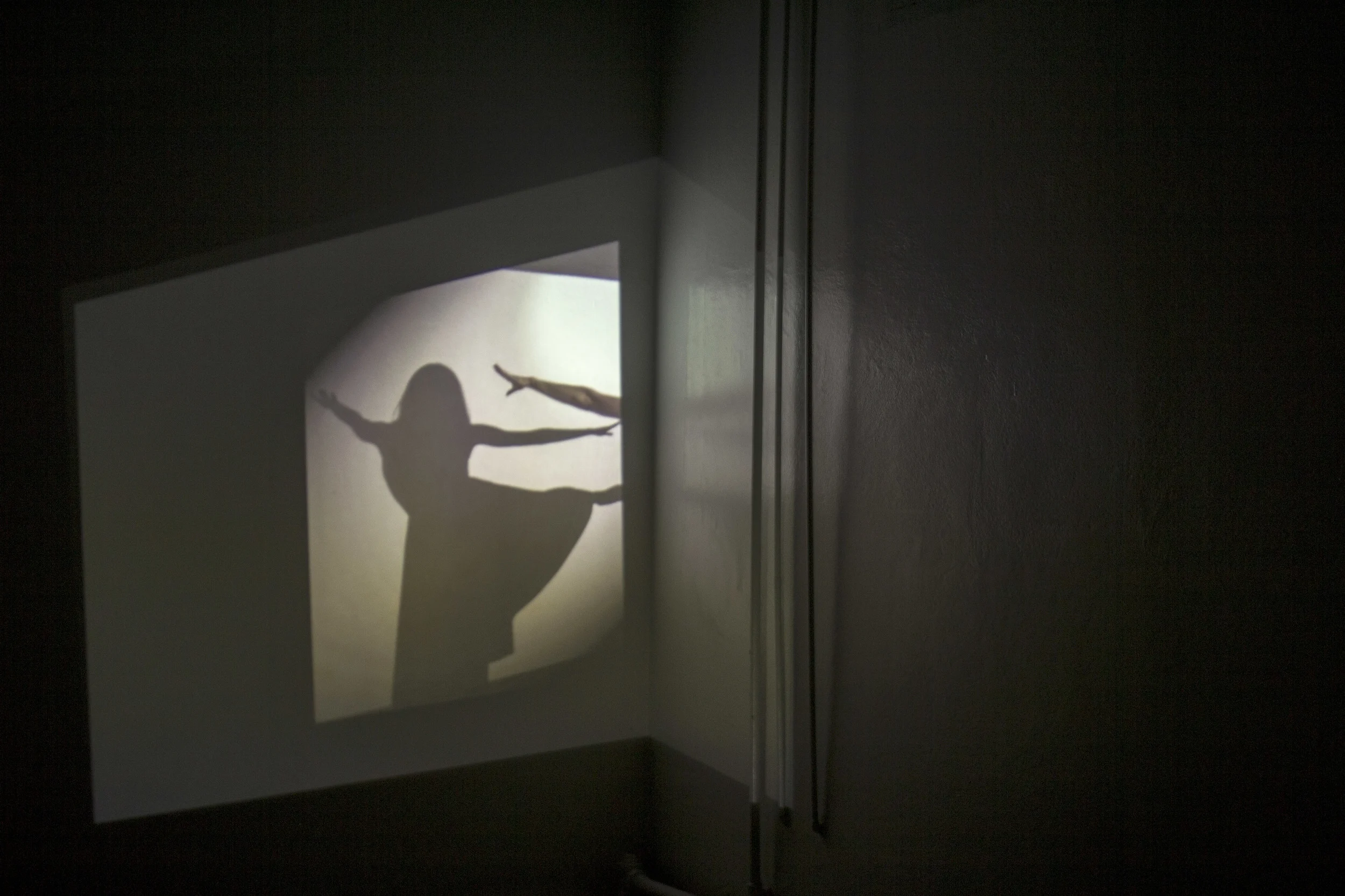 Across an expanse, Sarah O Donnell’s piece pitches across two rooms. In the first, a cord connects a mirrored projection of a dancer to a hidden sound in the next room. As the mirror spins at the end of a line, it throws the projected dancer’s image around the room. She flashes over the walls; the sound laments across the hall. Even when the sound’s source is found inside a suspended bucket held by a line of rope, it remains elusive, mournful. Here, all that is torn asunder is brought not into union, but into proximity, just touching. 