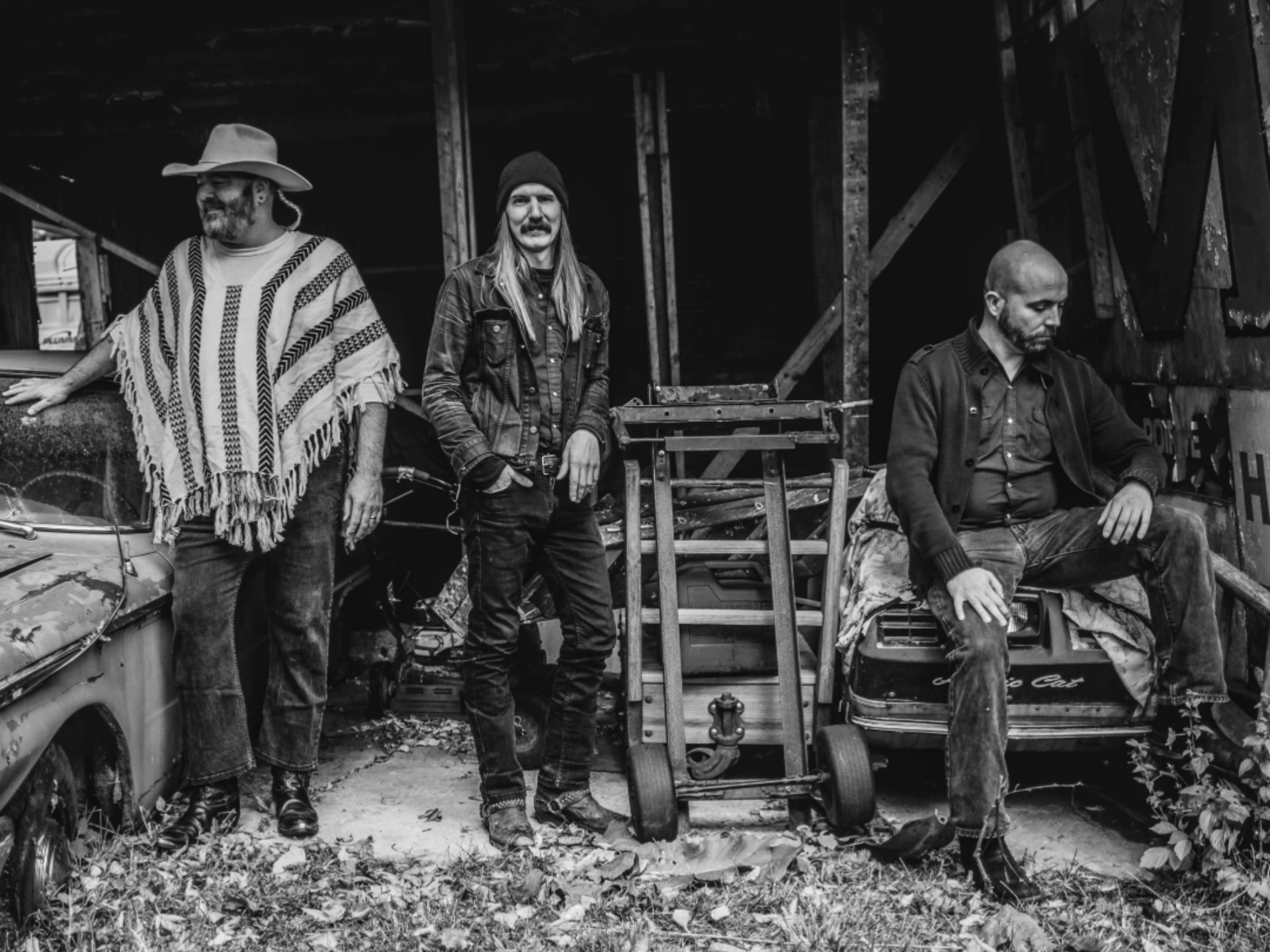 Three men standing and sitting in an abandoned, rustic setting with old cars and wooden structures around. The man on the left wears a large hat and striped poncho, the middle man has long hair, a beanie, and denim jacket, and the man on the right sits with one knee up, wearing a dark jacket and jeans, all in black and white.
