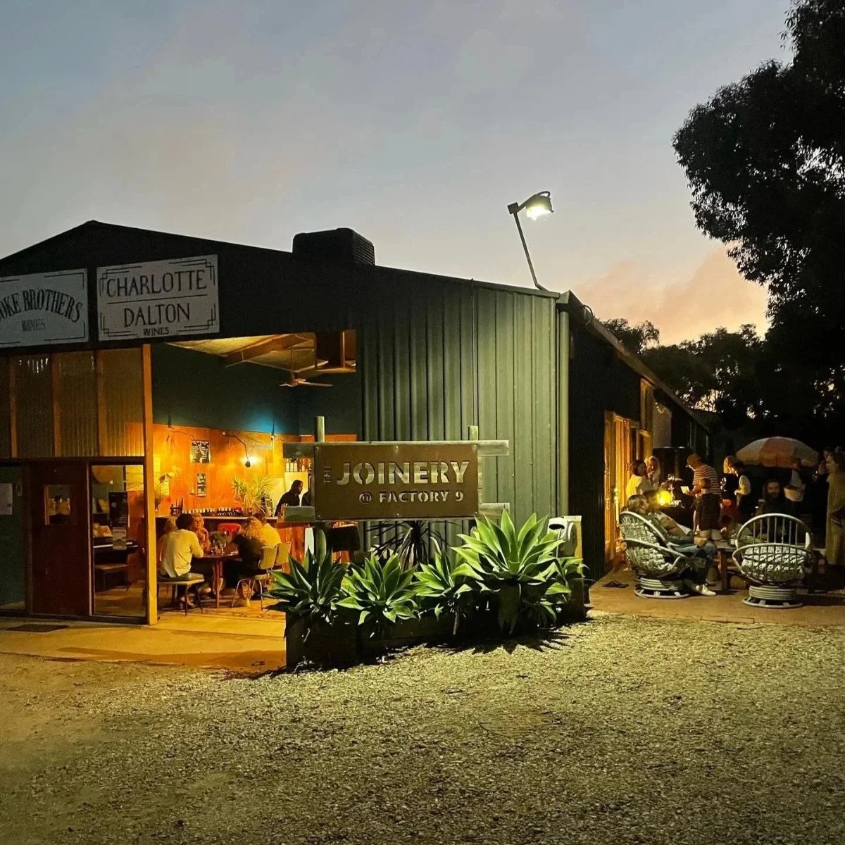 Exterior of The Joinery shed at sunset, groups of people are inside and outside drinking