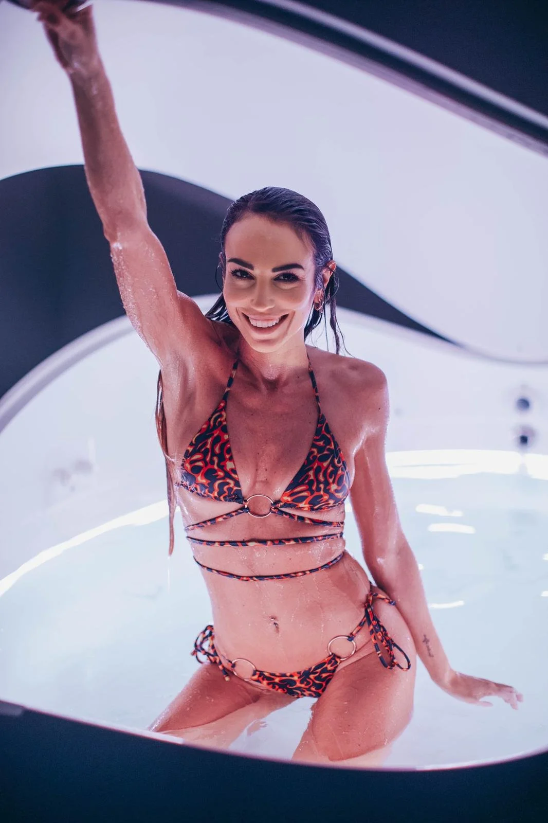 Una donna sorridente in costume da bagno a motivi rossi e blu, che si trova in una vasca idromassaggio, con acqua che le scorre sulla pelle.