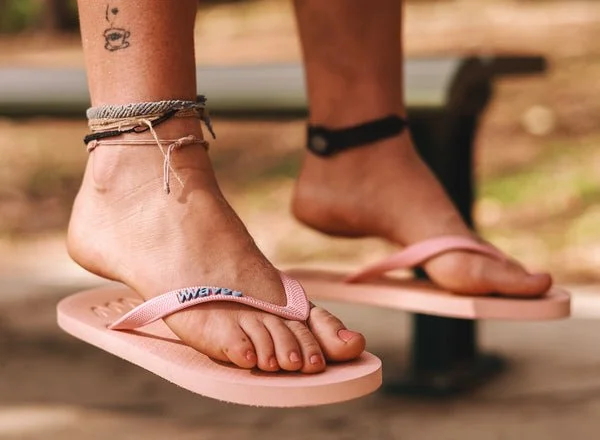 Person wearing flip-flops on a wooden bench, with visible tattoos and anklets.