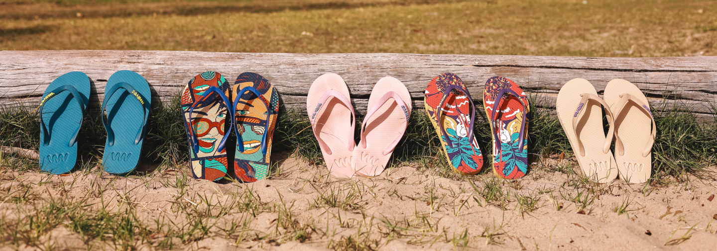 Six pairs of flip-flops in different colors and patterns placed on sand in front of a weathered wood boardwalk.