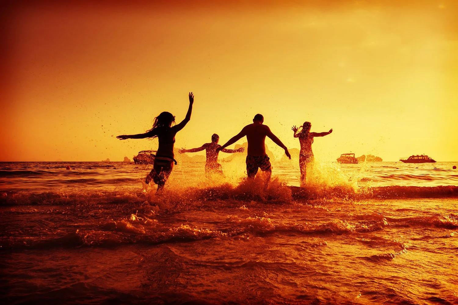 Silhouettes of four people running and playing in the ocean during sunset with boats in the background.
