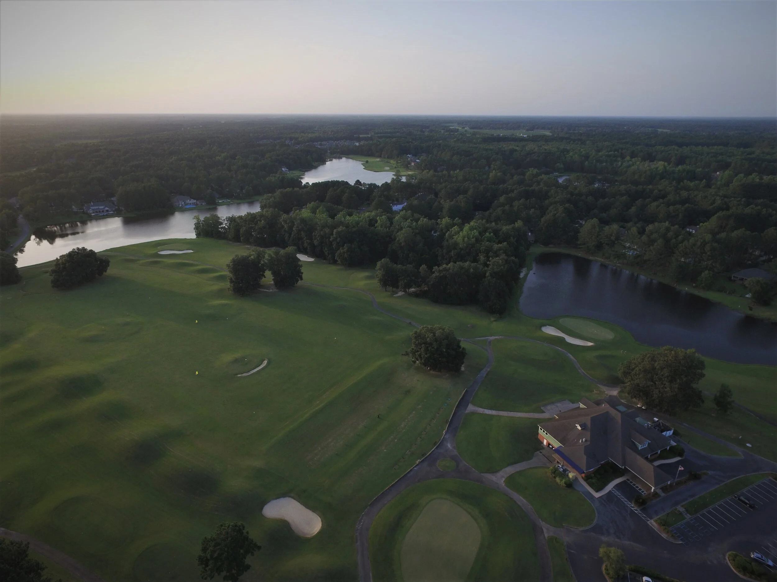 Aerial view of a golf course with green fairways, sand traps, and surrounding trees, lakes, and a building with parking lot.