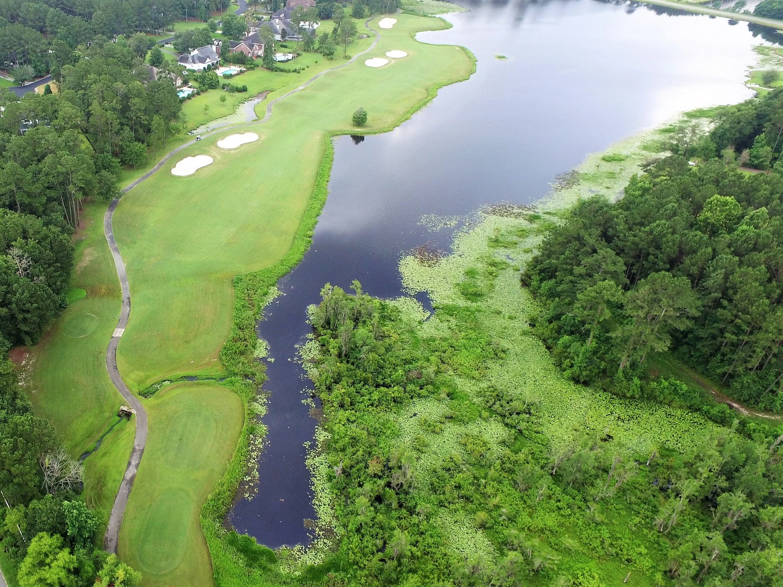 Aerial view of a golf course with a large water hazard surrounded by trees and a residential area in the background.