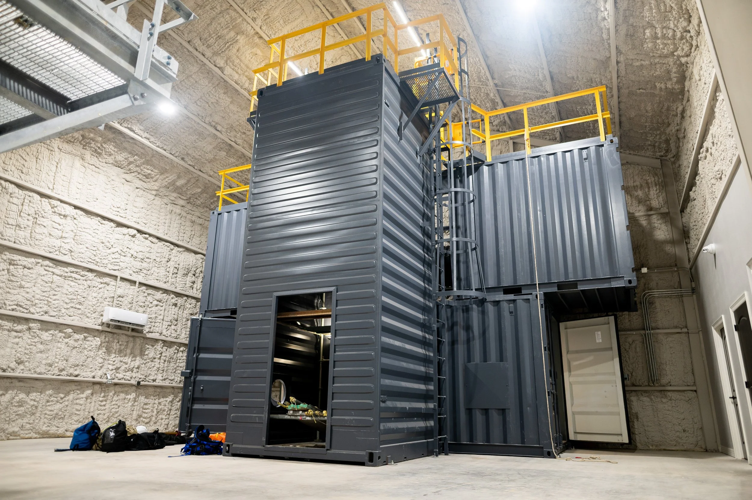 Large industrial container structure made from repurposed shipping containers, with yellow safety railings on top, situated inside a high-ceilinged industrial space.