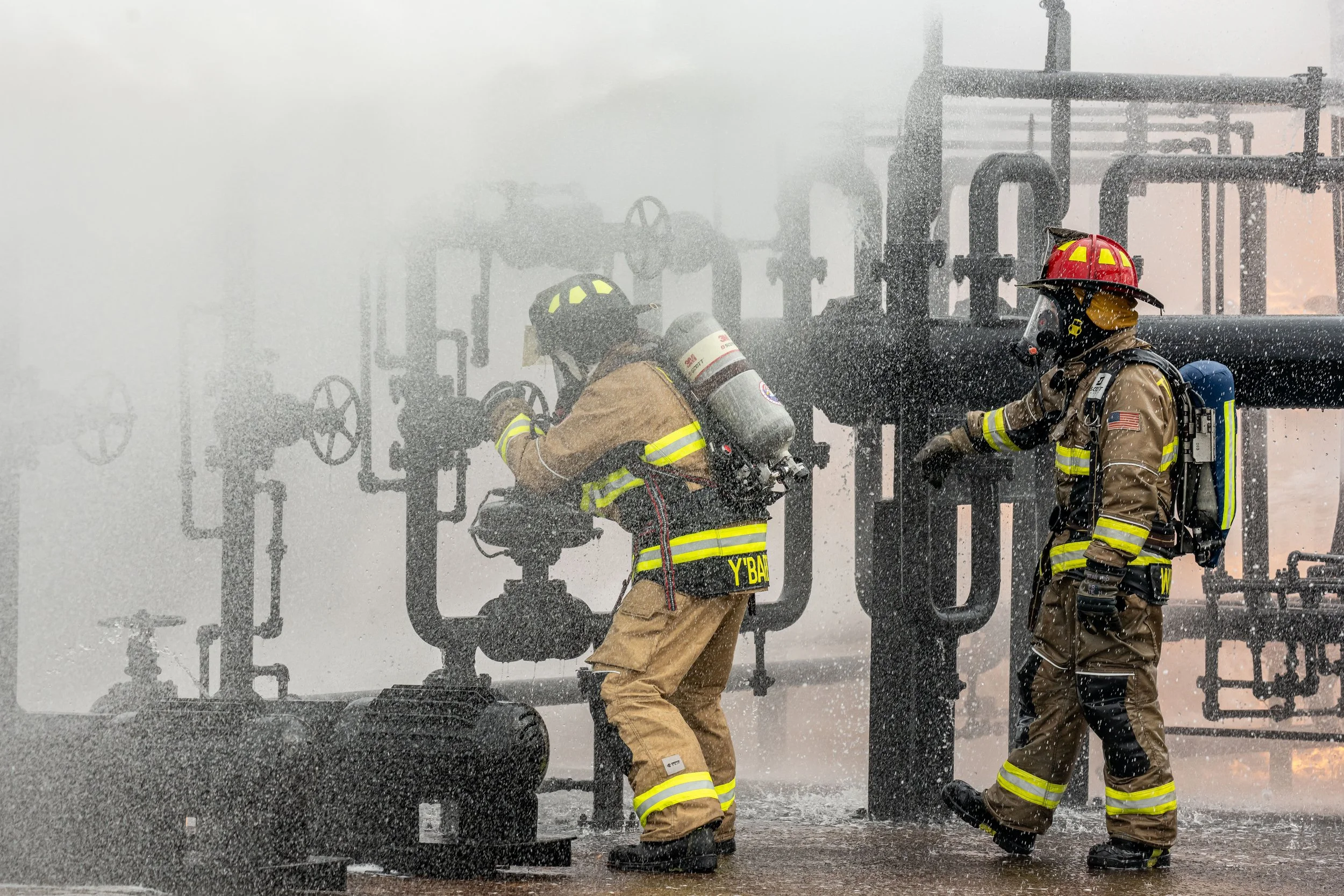 Two firefighters in protective gear working amidst spray and smoke at an industrial site with pipes and valves.