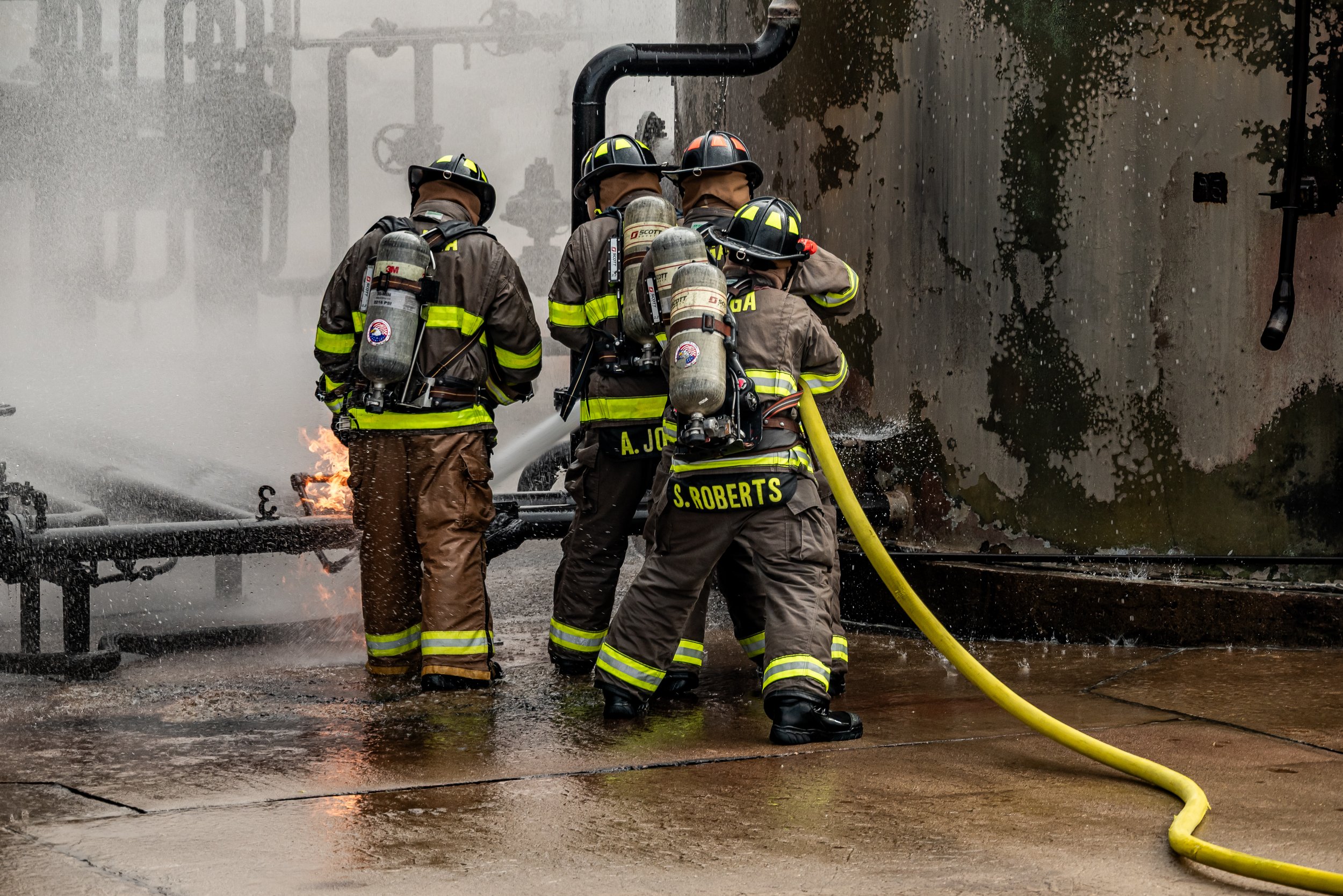 Firefighters operating a hose to put out a fire, with water spraying and flames visible on a surface.