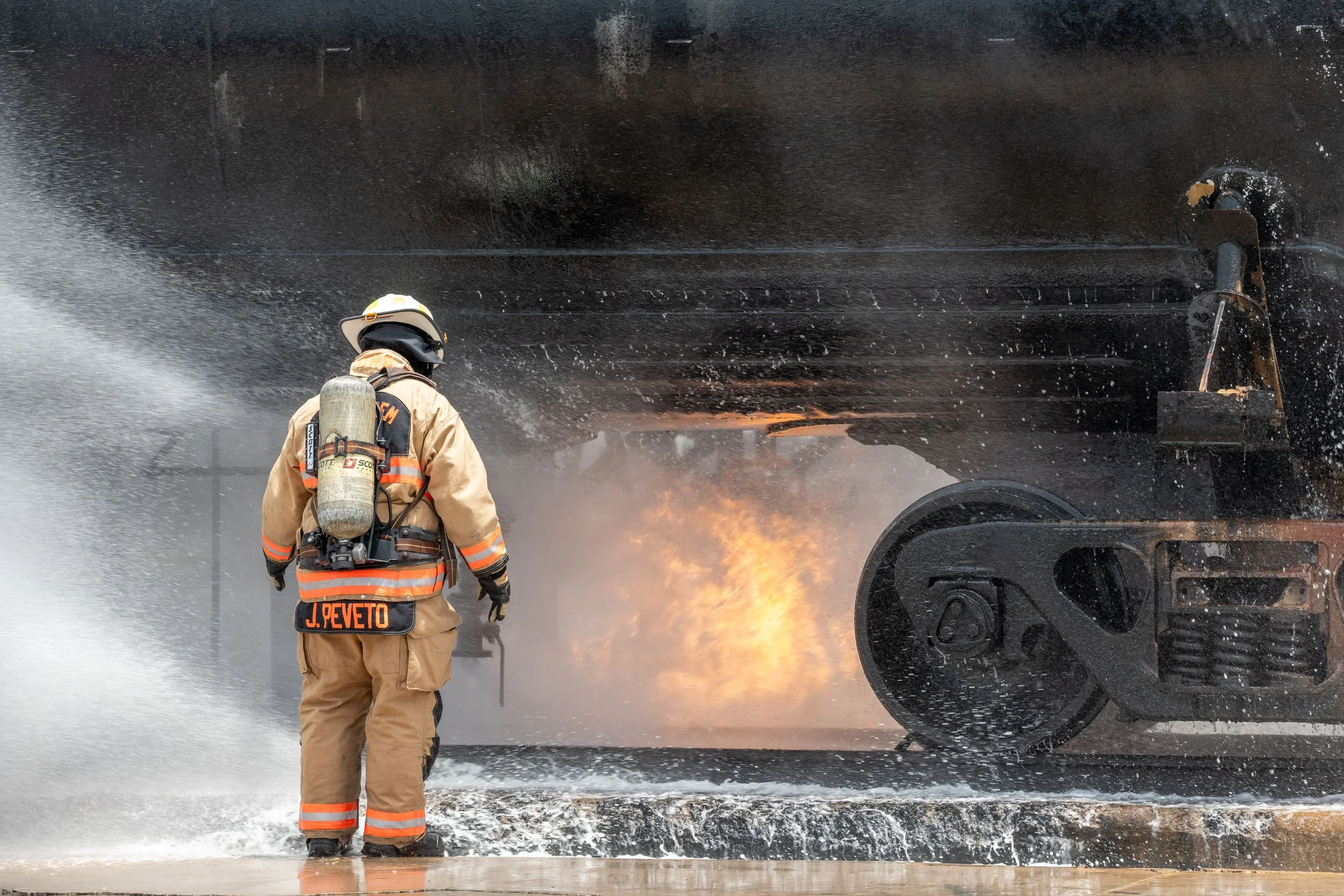 Firefighter in protective gear fighting fire on a large vessel with visible flames and water spray.