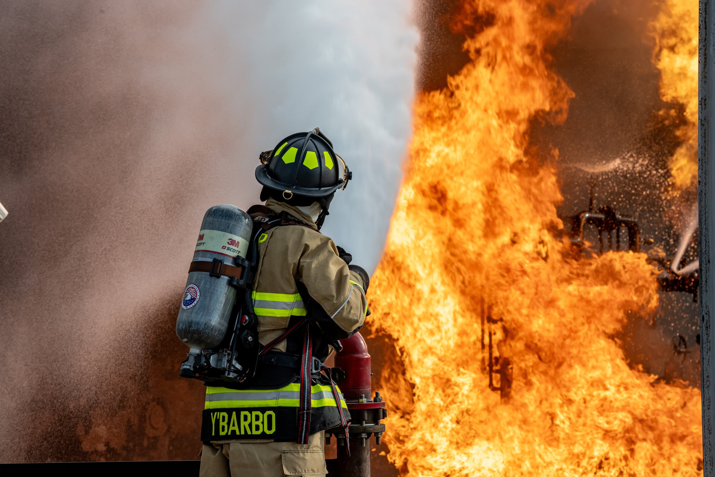 Firefighter in protective gear and breathing apparatus fighting a large blaze with flames and smoke.