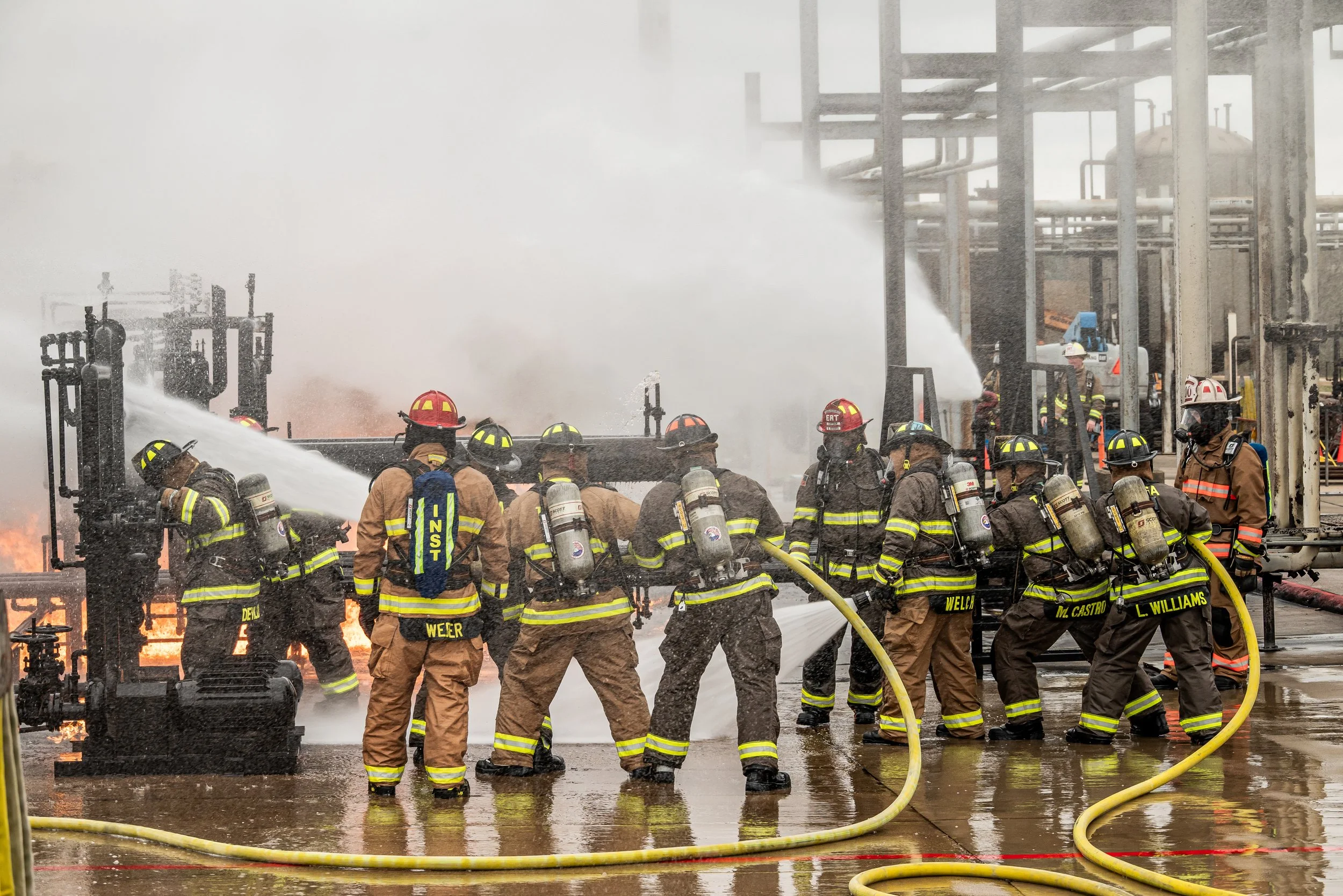 Firefighters working together to extinguish a fire at an industrial site, spraying water on flames amidst smoke and steel structures.