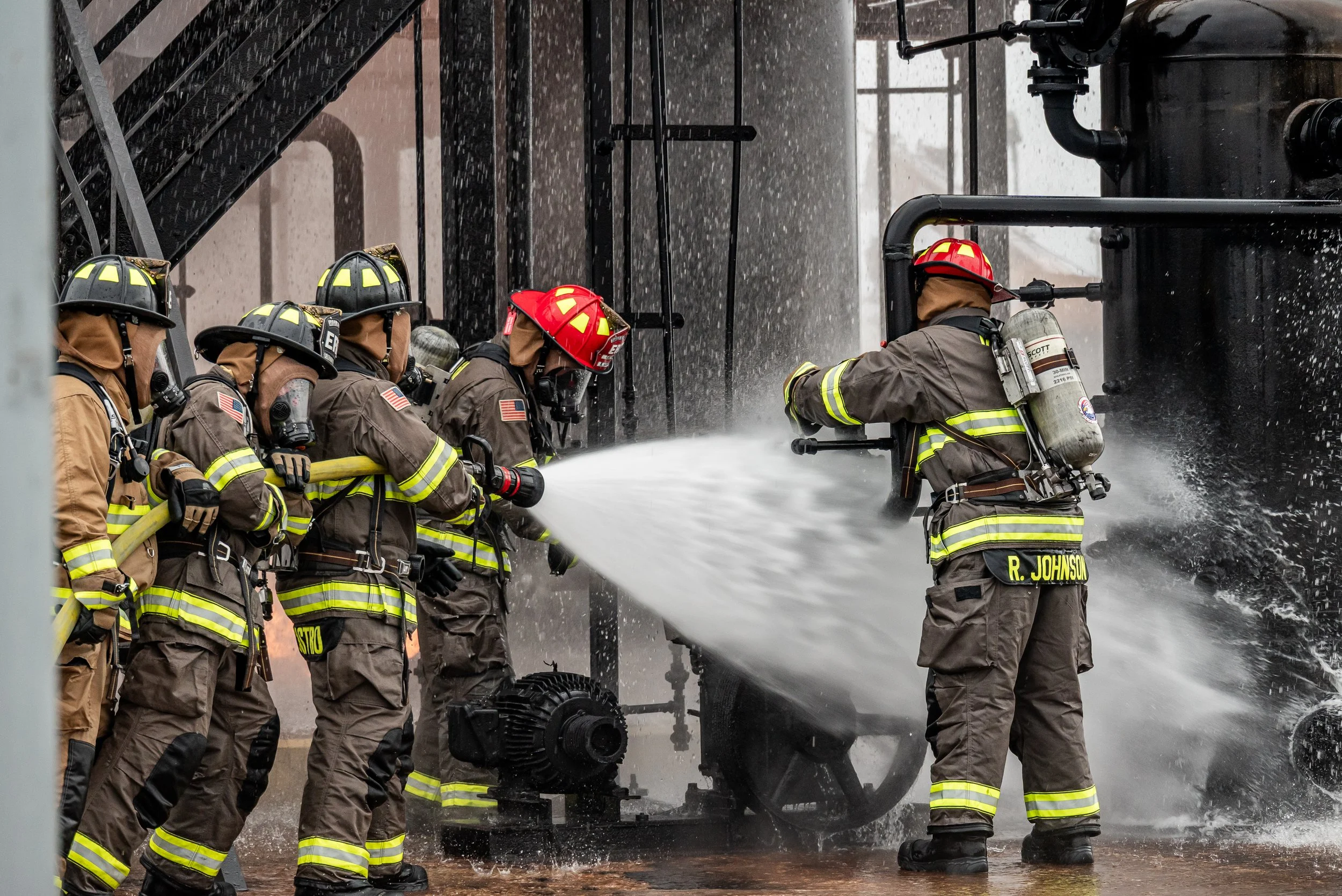 Firefighters in full gear spray water from a hose onto a fire, with water splashing and sprinklers overhead.
