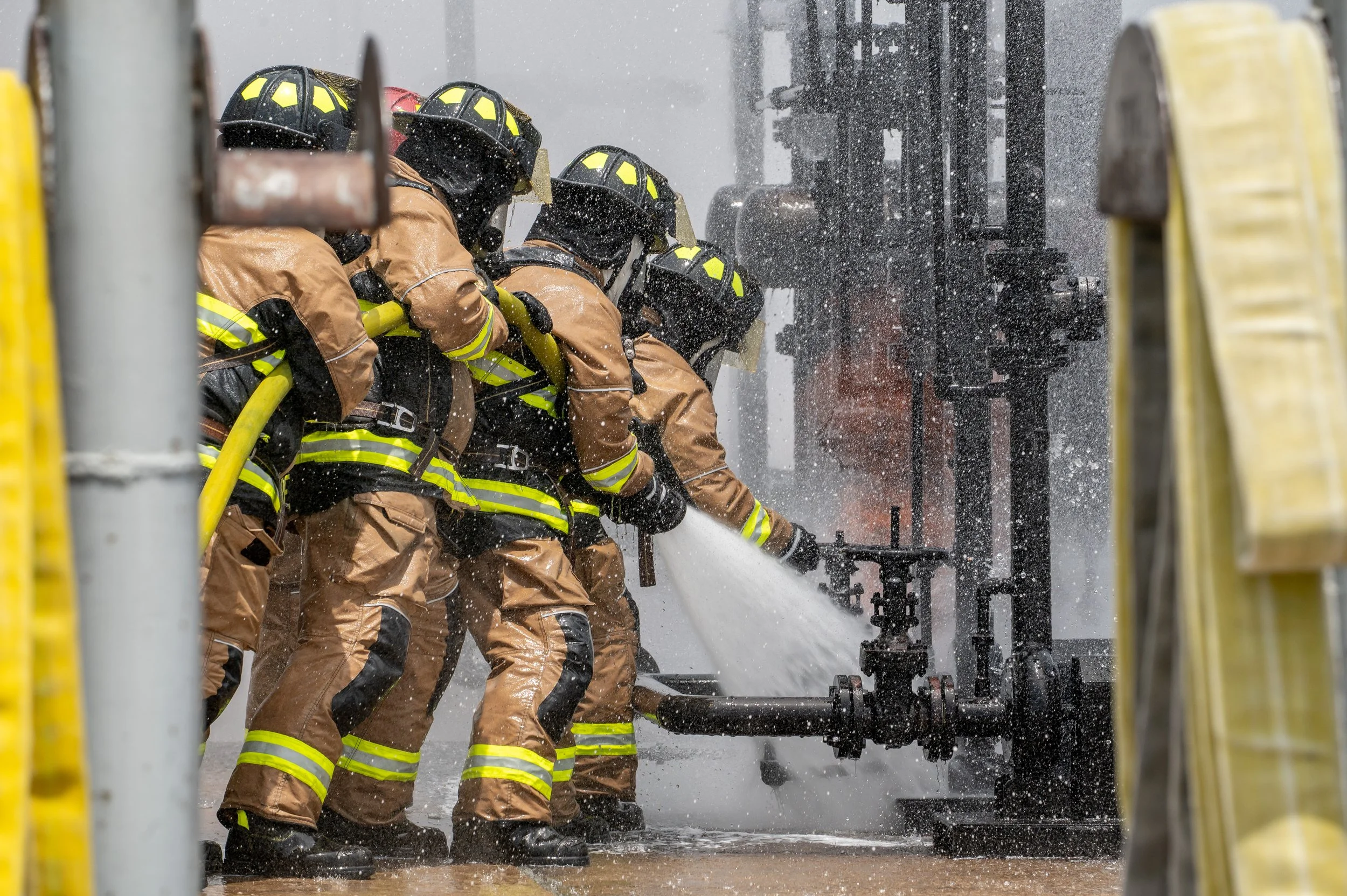 Firefighters in firefighting gear spray water onto a fire, working together to extinguish the flames.