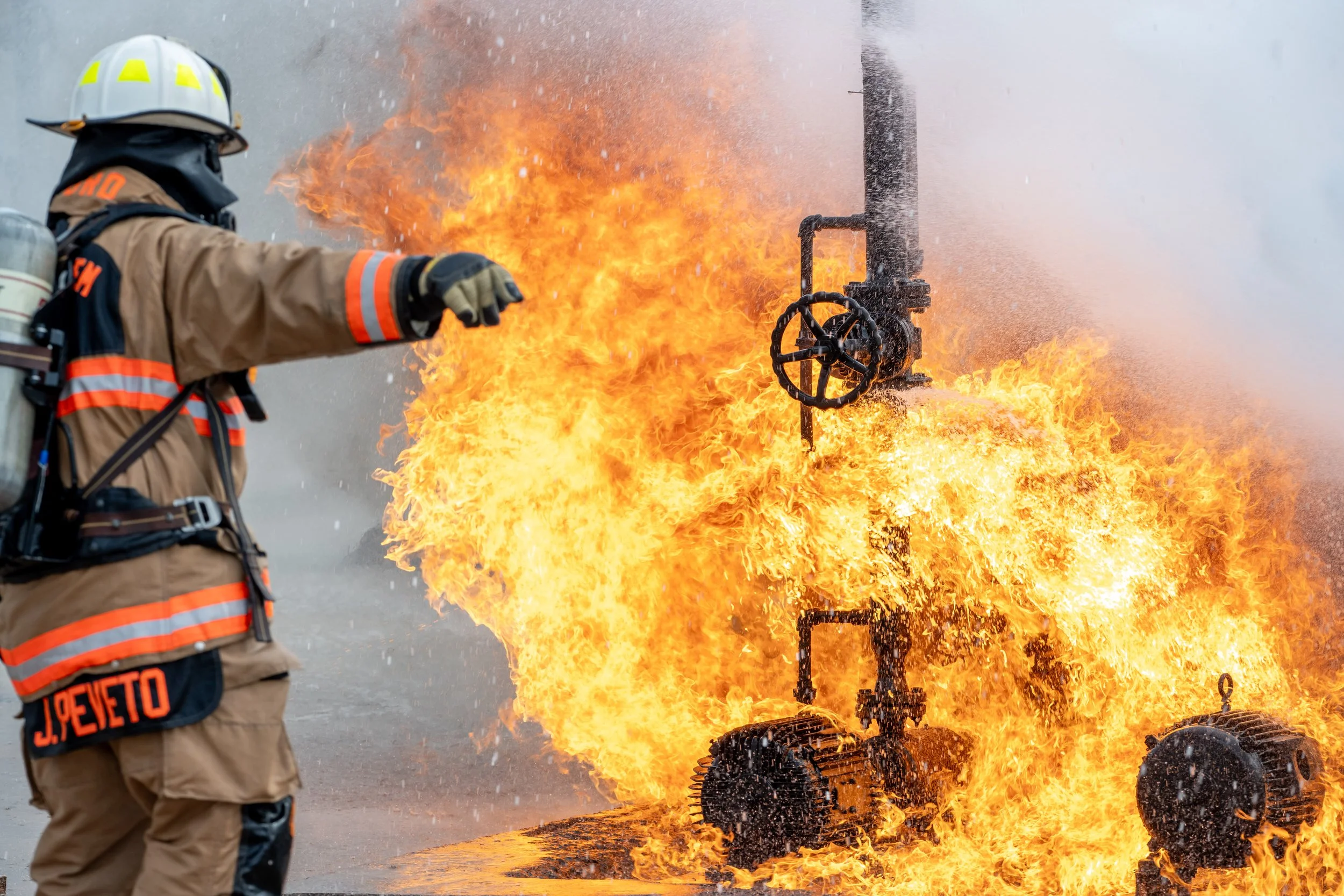 Firefighter in protective gear battling a large fire involving a burning pipeline or industrial equipment.
