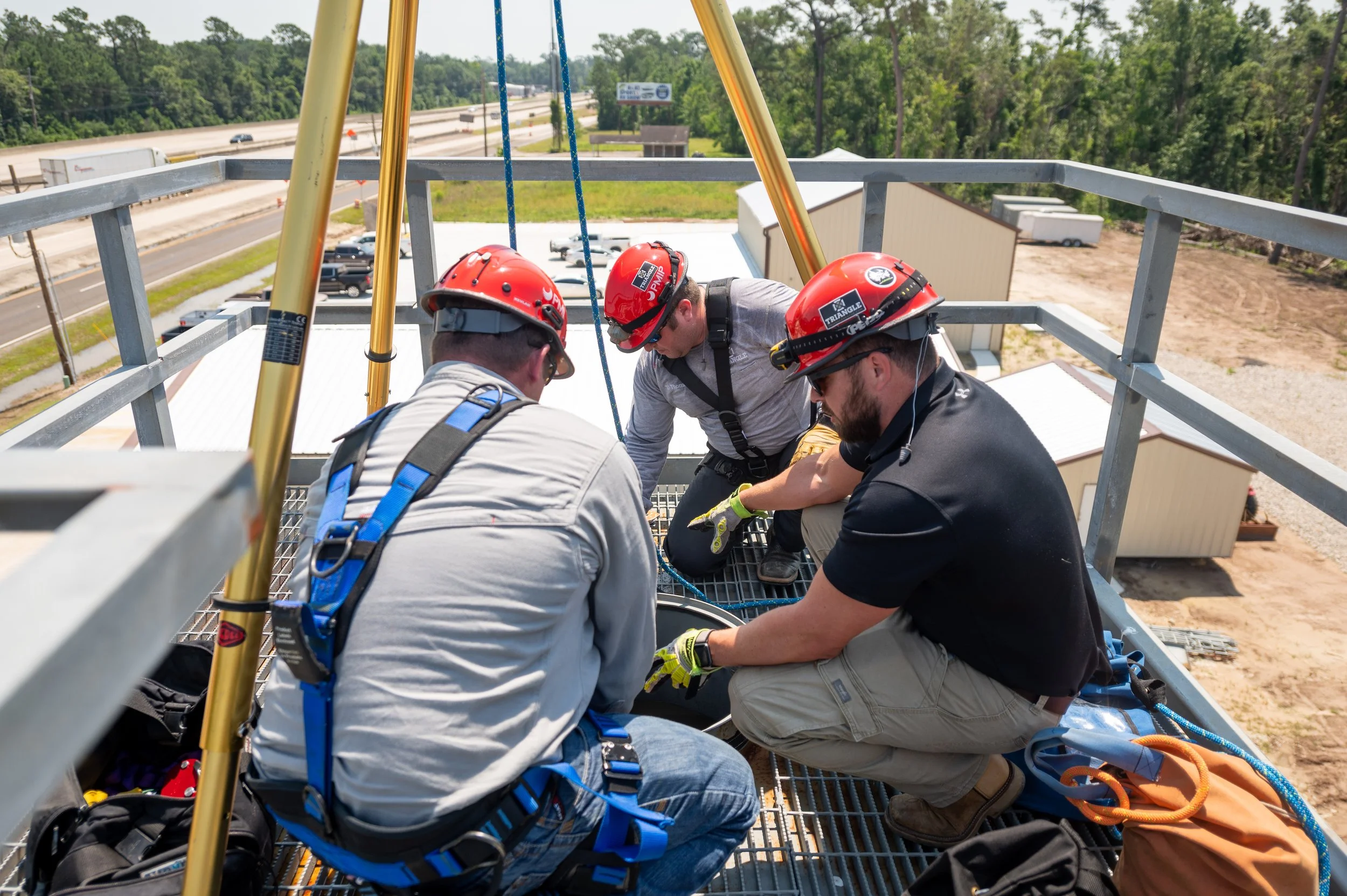 Three rescue workers in helmets and harnesses working together on a metal platform high above the ground, near a highway with vehicles passing by in the background.