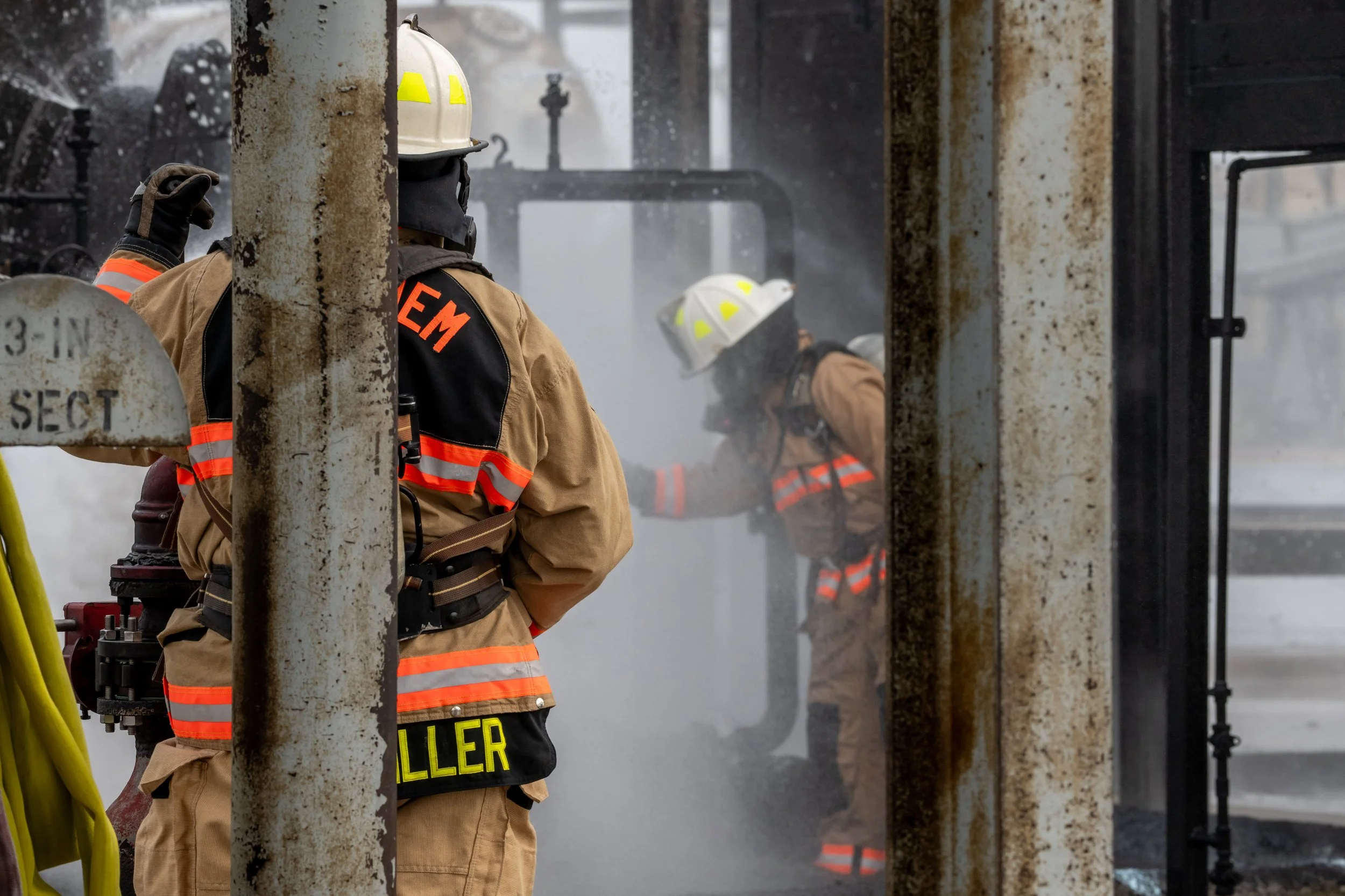 Firefighters in protective gear and helmets working amidst smoke and burnt equipment, responding to an emergency at a construction or industrial site.