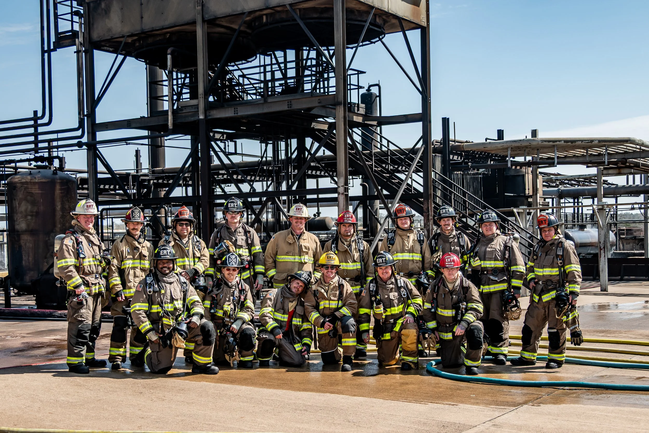 A group of firefighters in firefighting gear posing in front of an industrial fire site, with pipes and metal structures behind them.