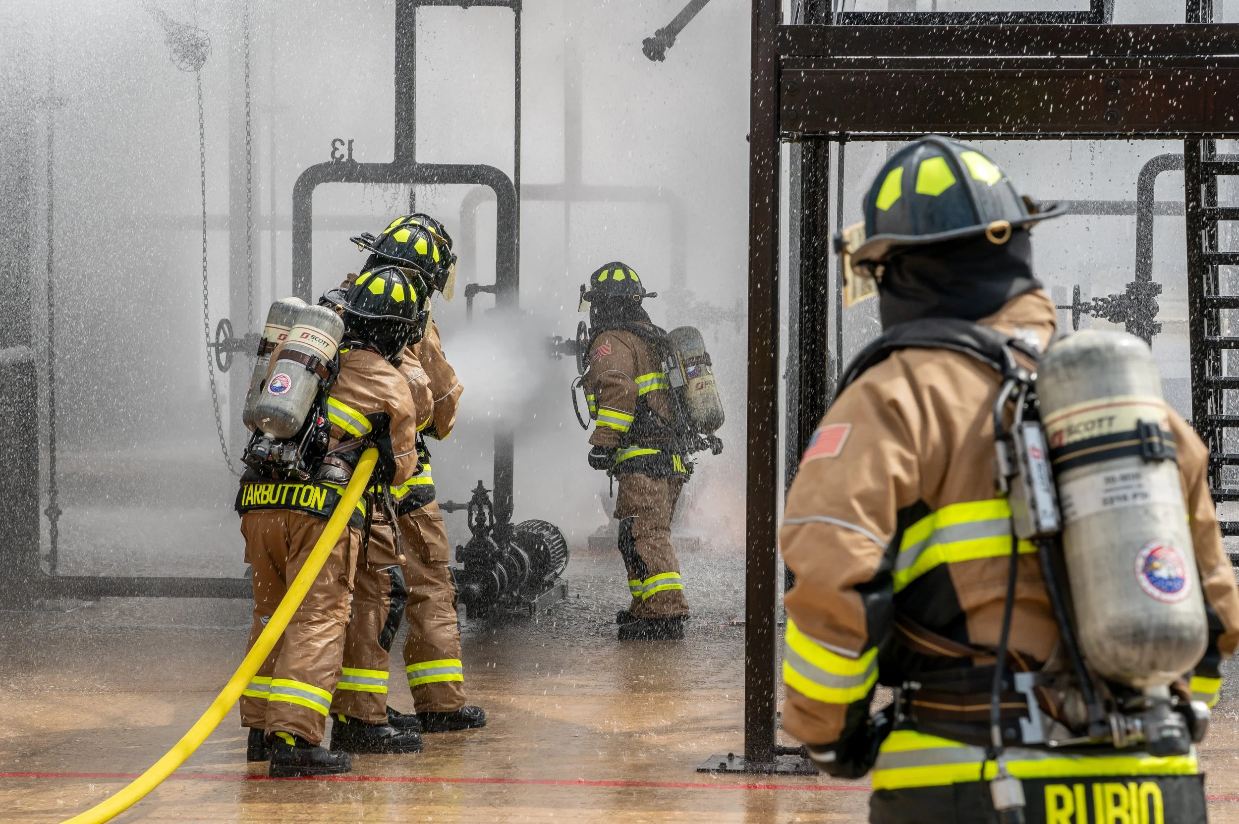 Firefighters in firefighting gear hosing down a fire, with water and smoke around them.