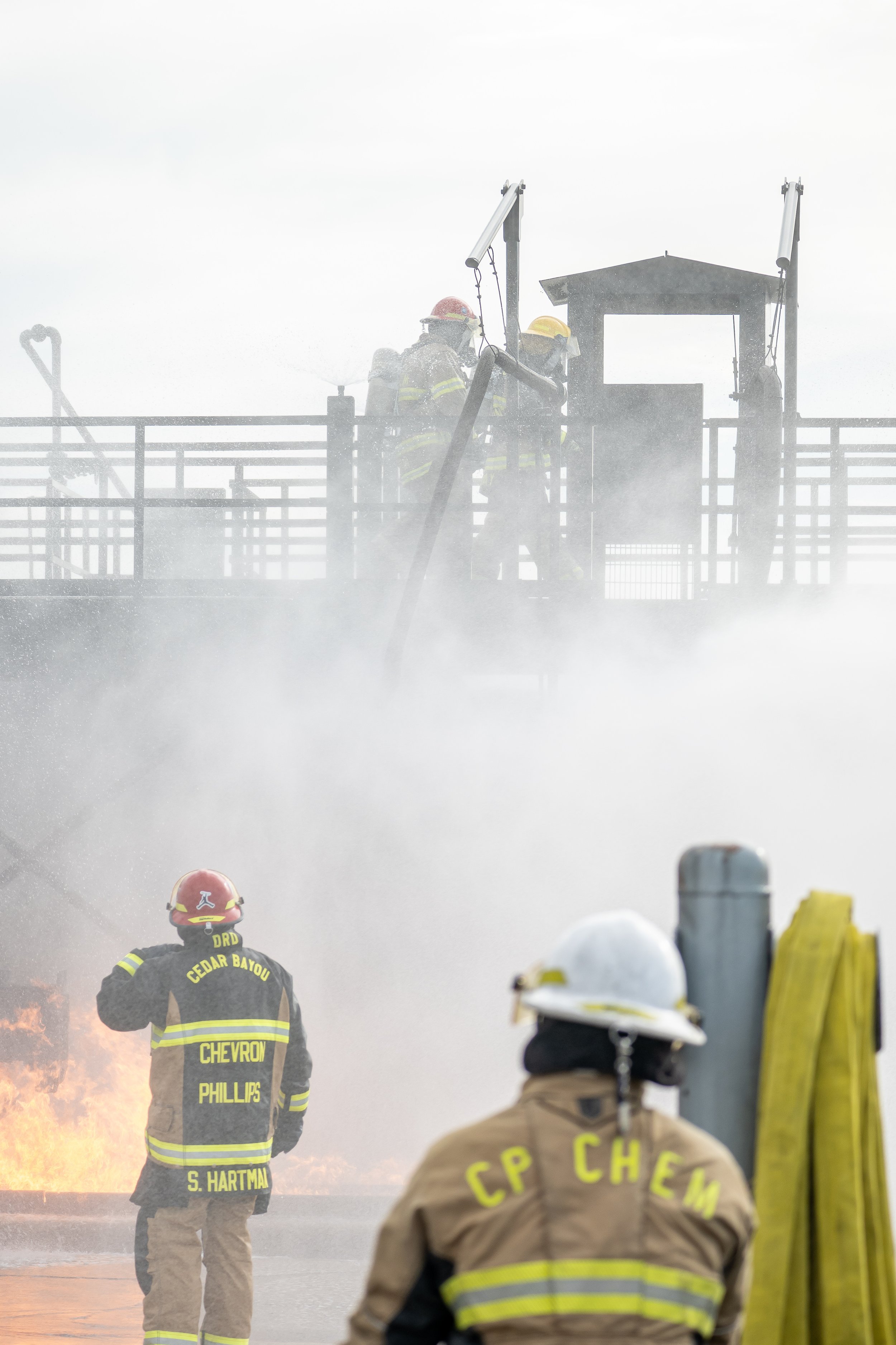 Firefighters in protective gear respond to a fire, with thick smoke and flames visible, on a boardwalk or similar structure.