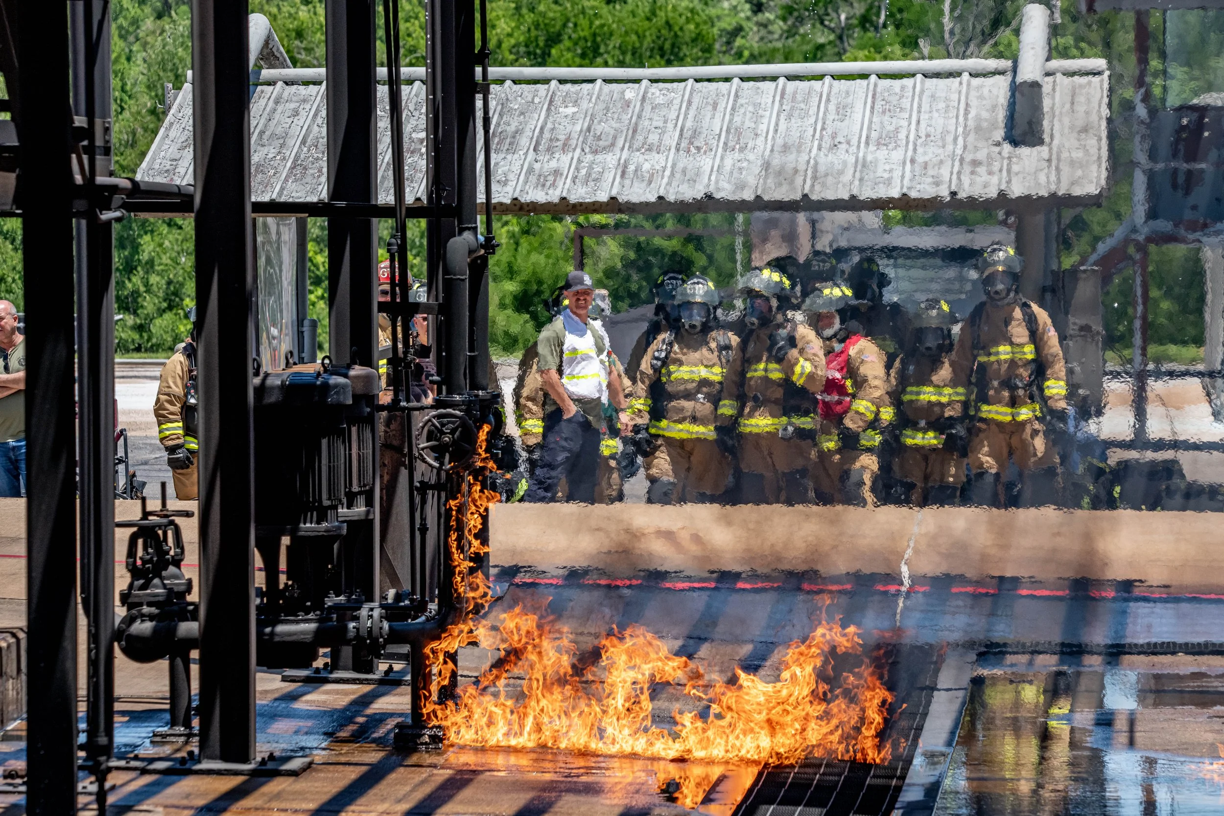 Firefighters in full gear standing behind a blaze on a roof, with flames and smoke visible, during a training or emergency response.