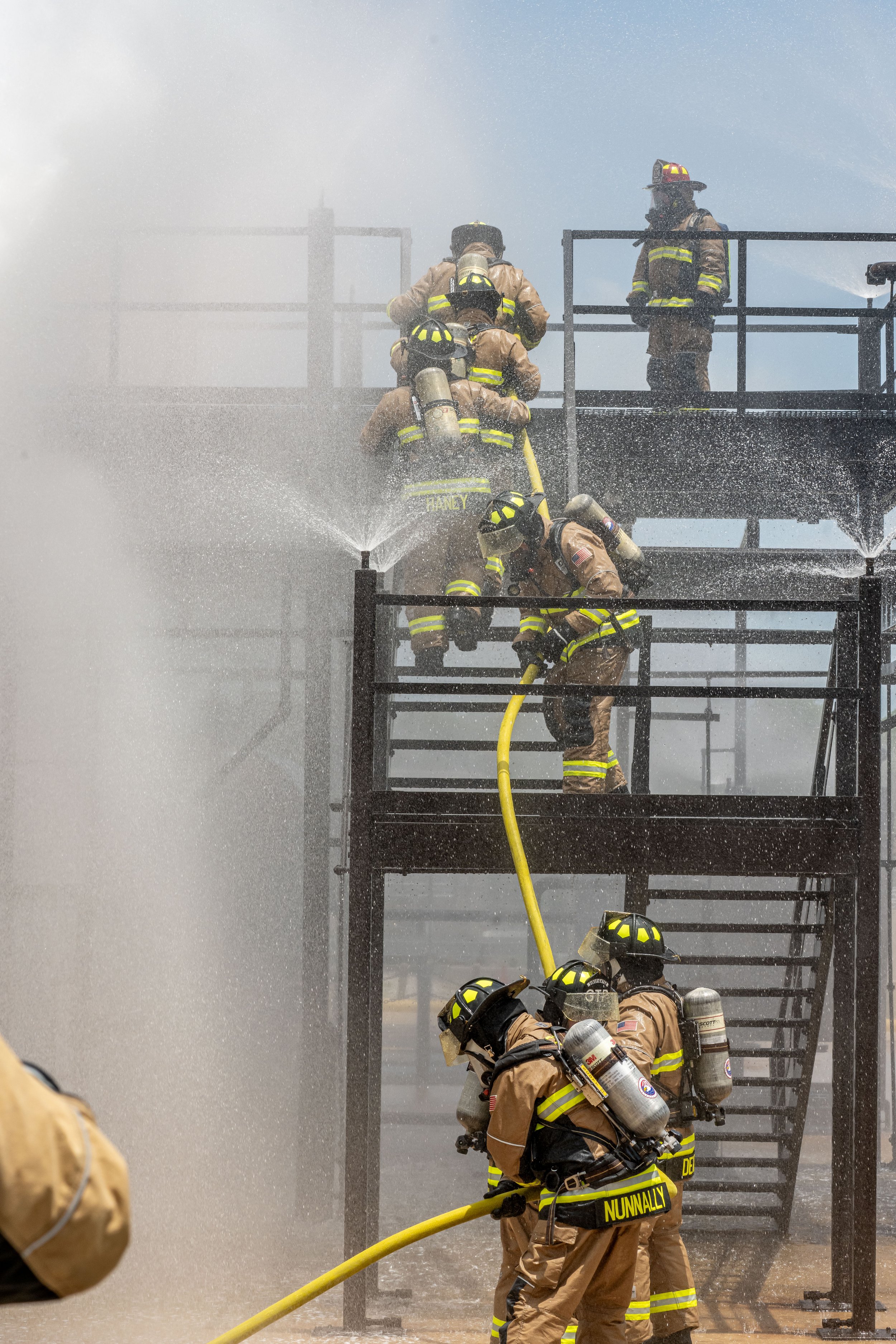 Firefighters in tan uniforms with yellow reflective strips and helmets are fighting a fire on a multi-level metal structure, using hoses that spray water, with some standing on upper and lower levels, amidst a cloud of smoke and steam.